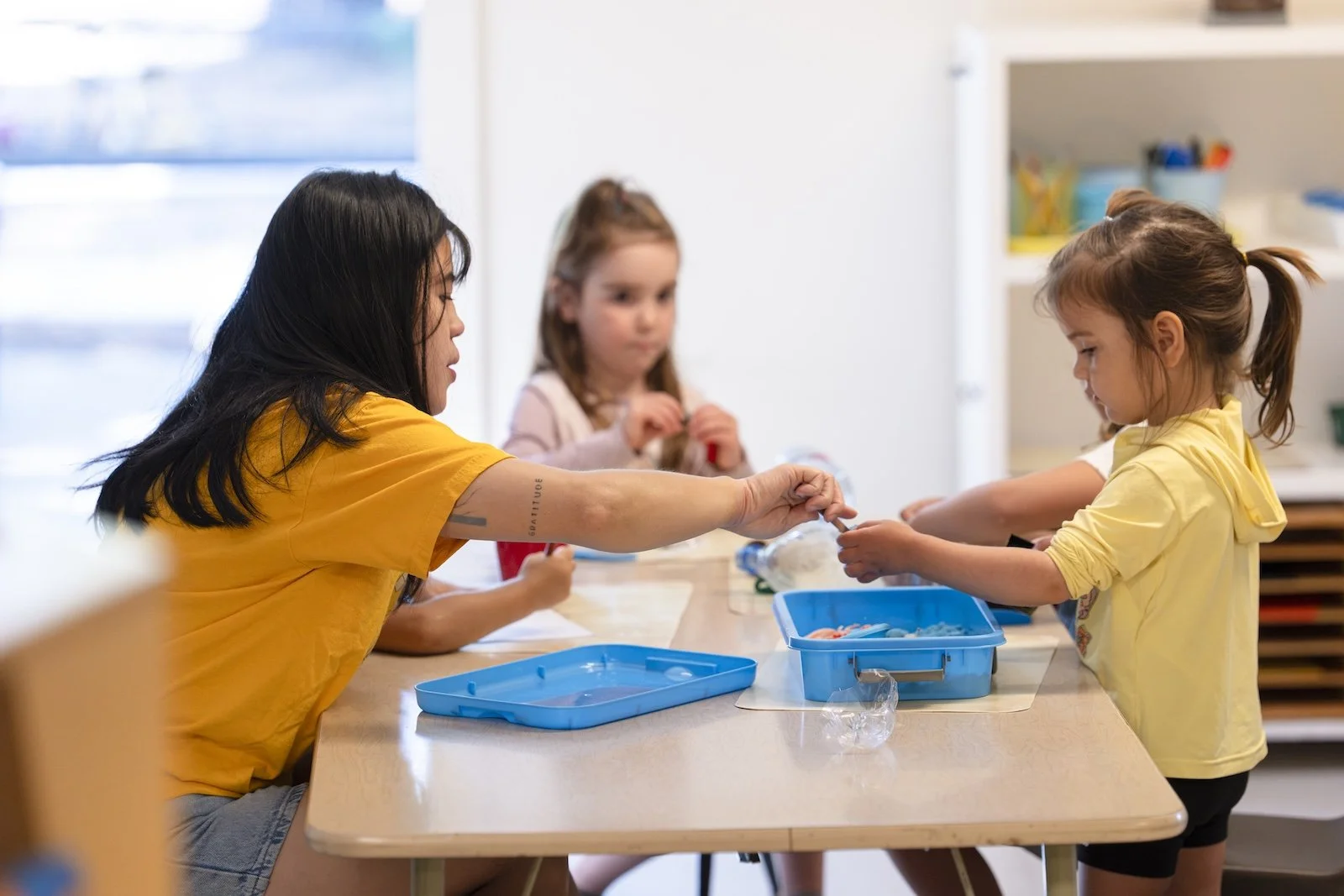 A woman and two young girls are sitting at a table, engaging in a craft activity. The woman in a yellow shirt is handing something to one of the girls, who is also wearing a yellow shirt. The girl in the background is focused on the activity. The table has blue containers and craft supplies.