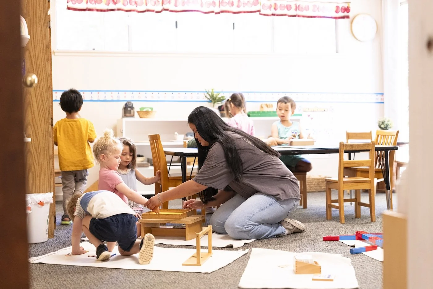 Teacher kneeling on the floor helping children with a wooden educational activity, classroom scene with children at a table and playing on the carpet.