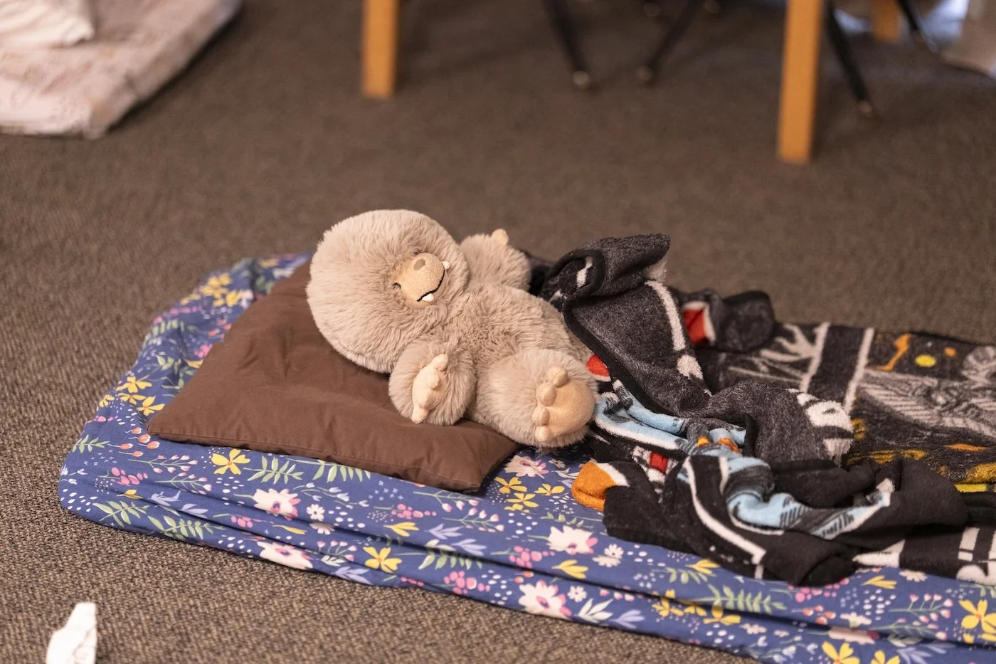 A plush sloth toy lying on a brown pillow on top of a blue floral-patterned mattress, partially covered with a black blanket with colorful designs, on a carpeted floor.