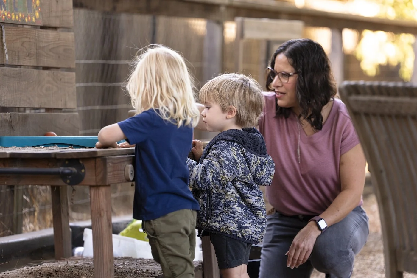A woman with glasses and dark hair kneeling next to two young boys at an outdoor wooden table, looking at something on the table.