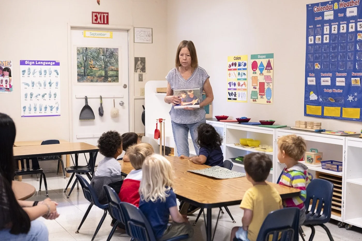 A teacher holding a book and standing in front of a group of young children seated at tables in a classroom.