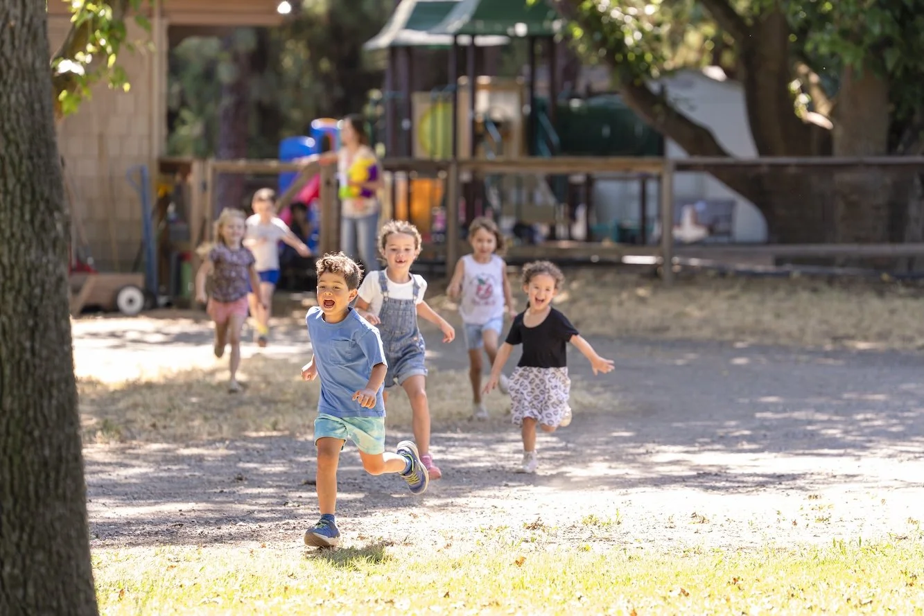 Children running and playing outdoors in a park or playground area, with trees and a fence in the background.