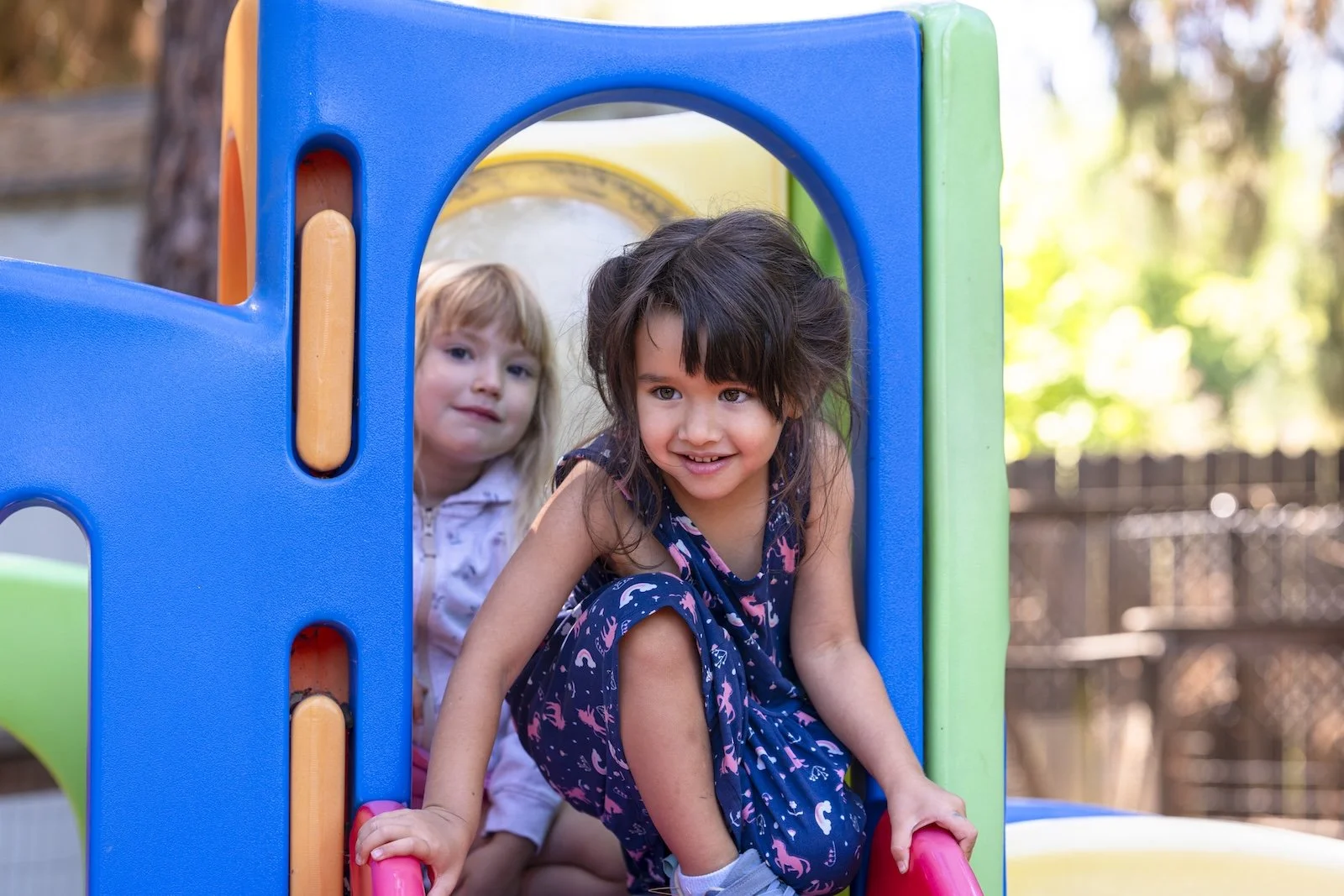 Two young girls playing on a colorful playground structure outdoors in a backyard, with trees and a fence in the background.
