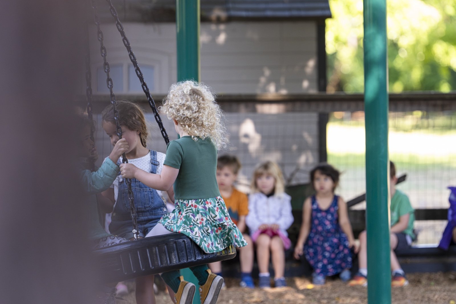 Children sitting on a swing and bench in a backyard or park, with two girls on the swing in the foreground, and four girls sitting on a bench in the background, near a gray house.