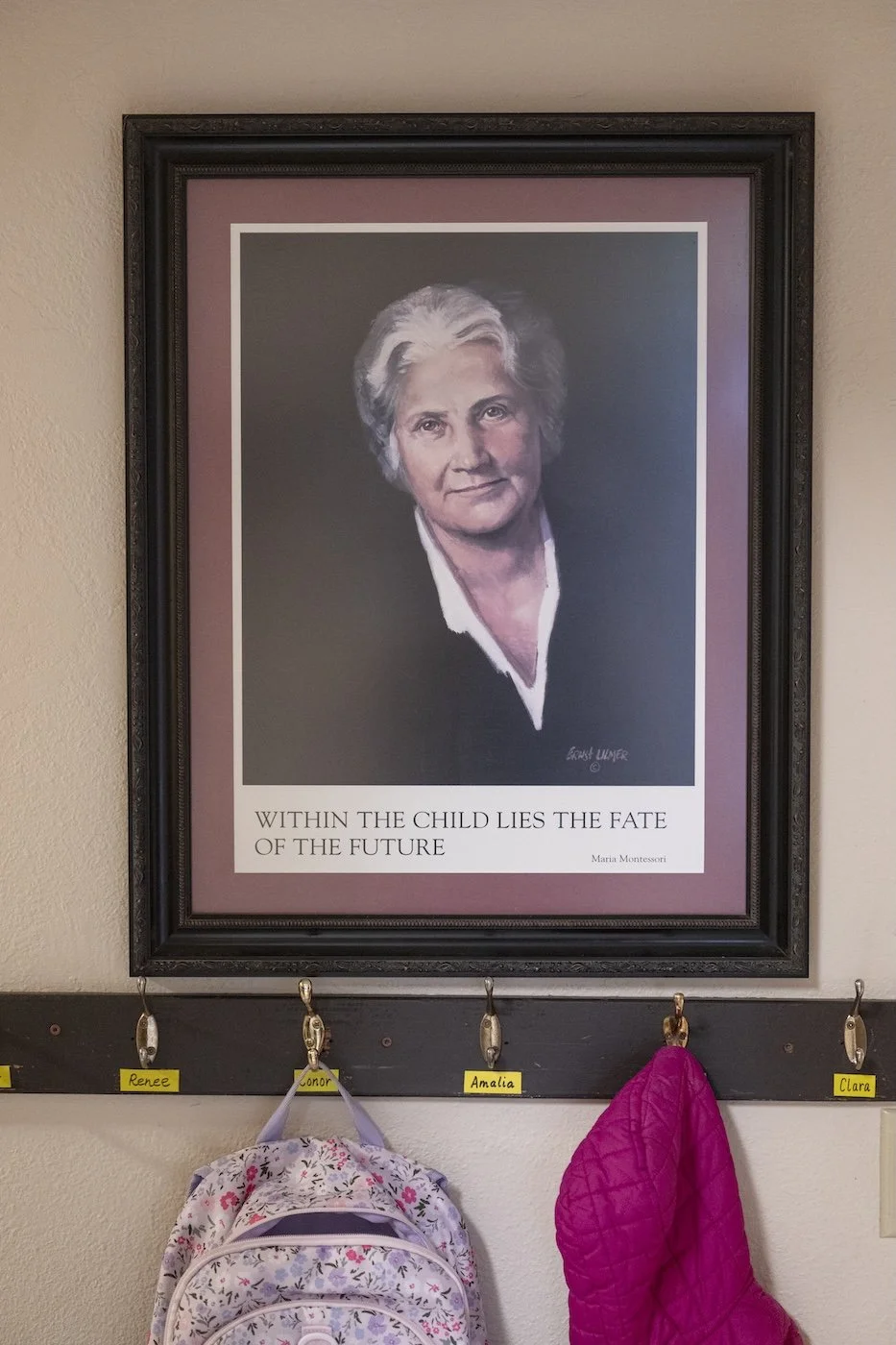 Framed portrait of Marie Montessori hanging on a wall. Below the frame are four hooks labeled with children's names, with backpacks hanging on two of them.