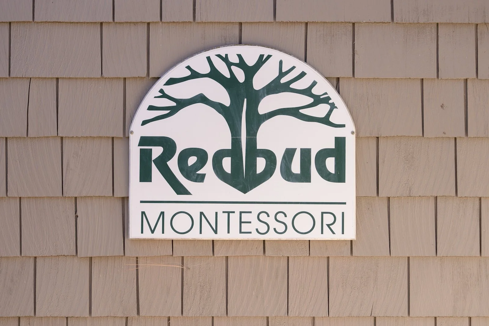 A sign on a beige wooden shingle wall that reads "Redbud Montessori" with a stylized tree logo.