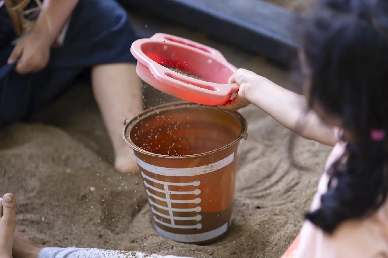 Child pouring sand into a large container with a smaller, pink sand sifter in a sandy outdoor area.