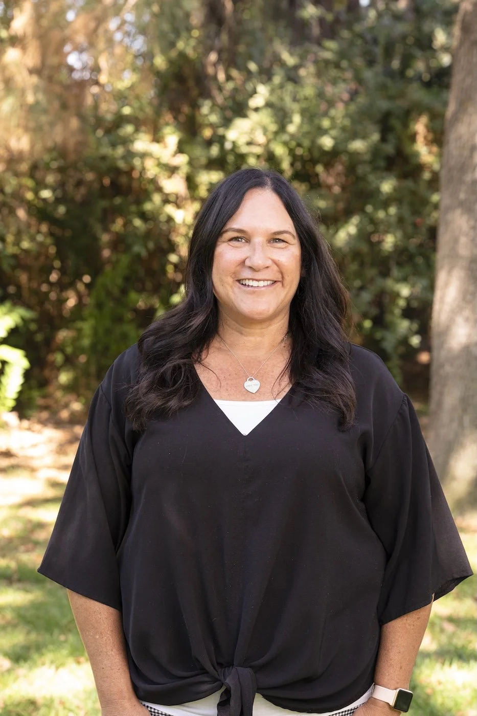 A woman with long dark hair smiling outdoors, wearing a black top with a front knot, a necklace, and a smartwatch, standing in front of trees and foliage.