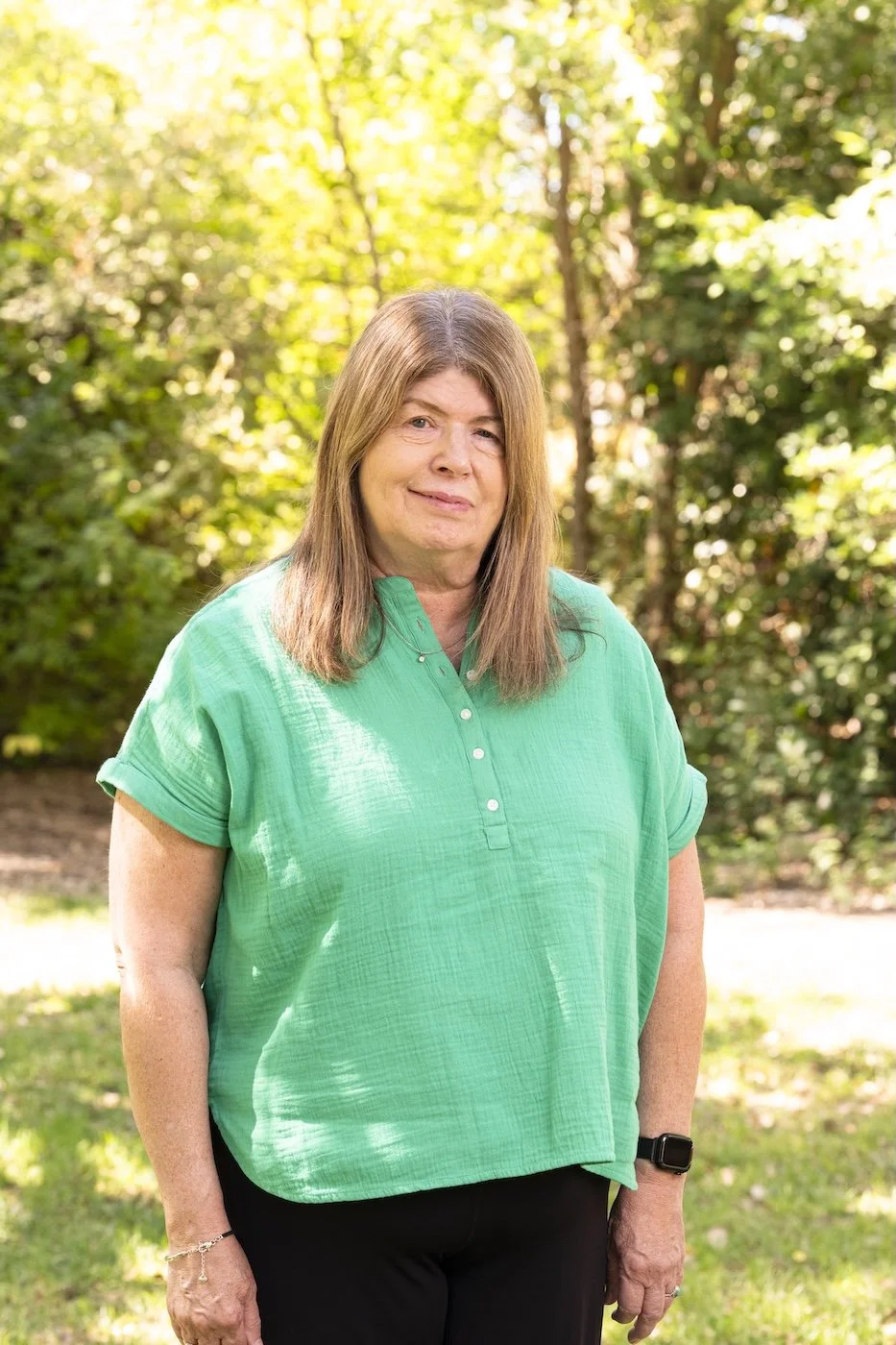A middle-aged woman with brown hair standing outdoors in a park, wearing a bright green blouse and black pants, with trees and sunlight in the background.