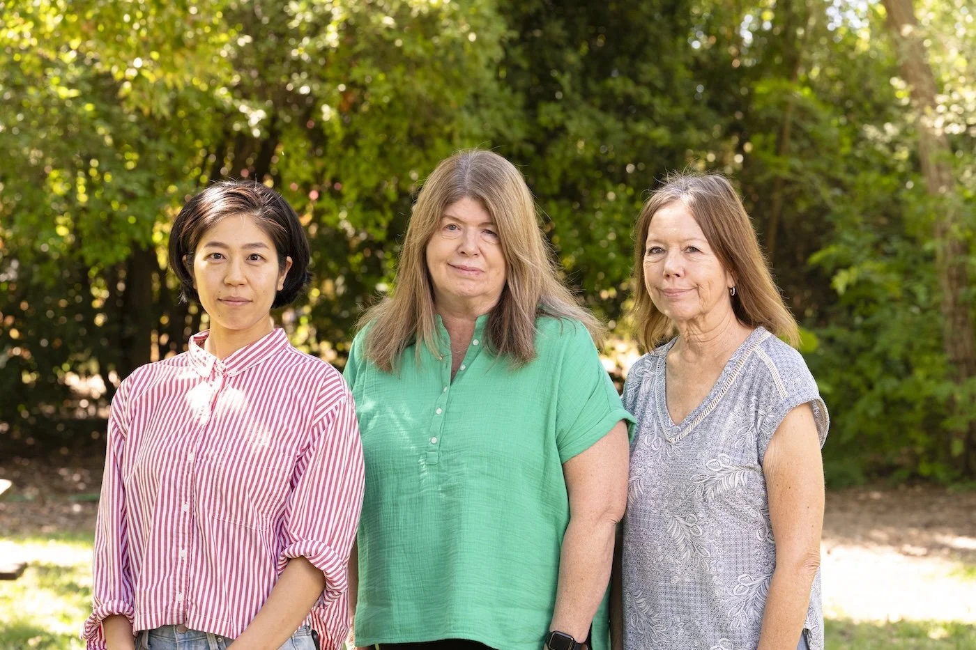 Three women standing outdoors in front of trees on a sunny day, smiling gently at the camera.