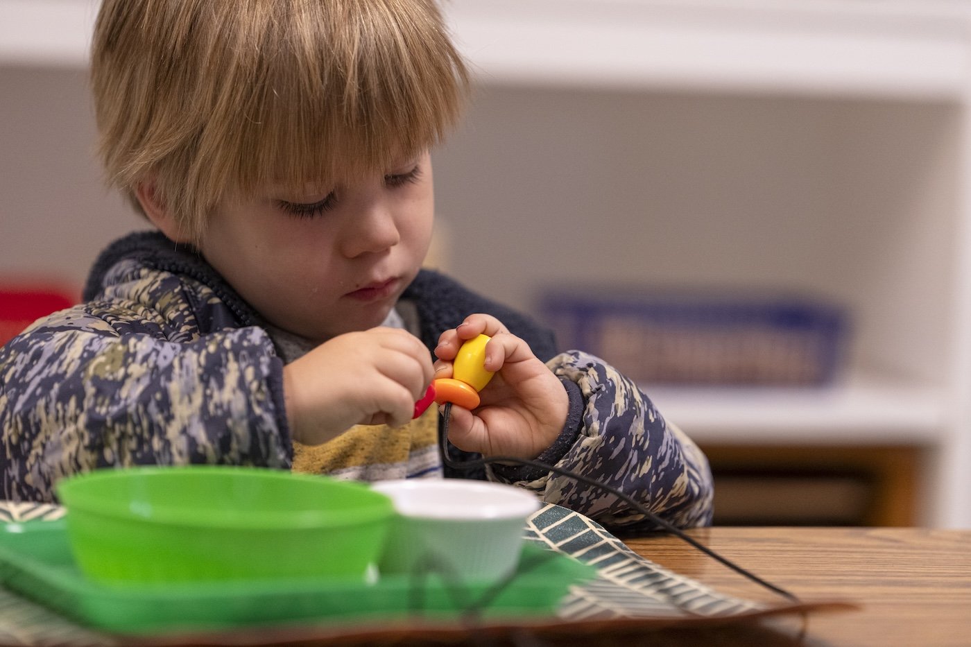 A young boy with blond hair concentrating on assembling a toy on a wooden table, with colorful plastic pieces and a green tray in front of him.
