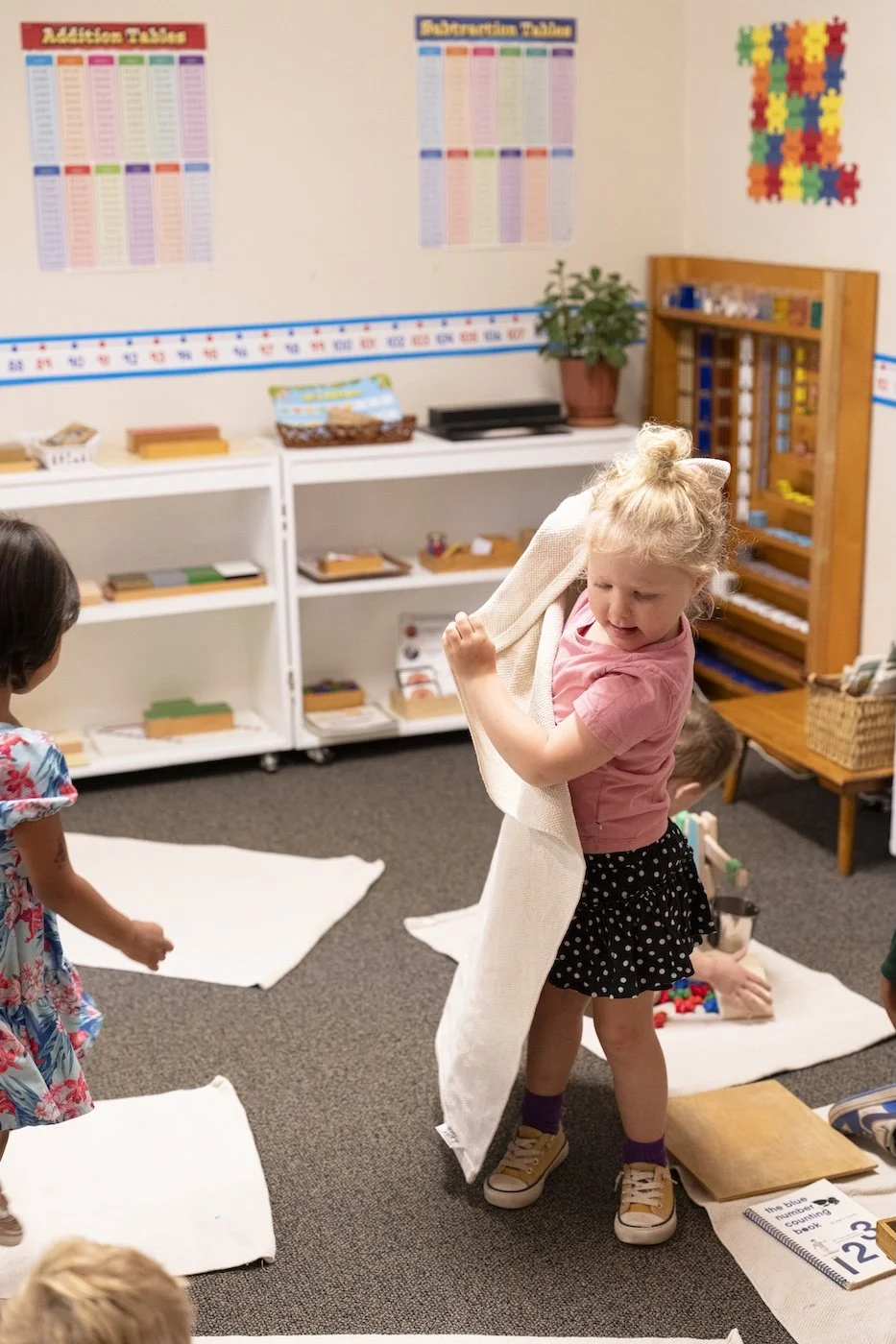 A young girl in a classroom holding a beige cloth or blanket over her shoulder while surrounded by other children and educational materials.
