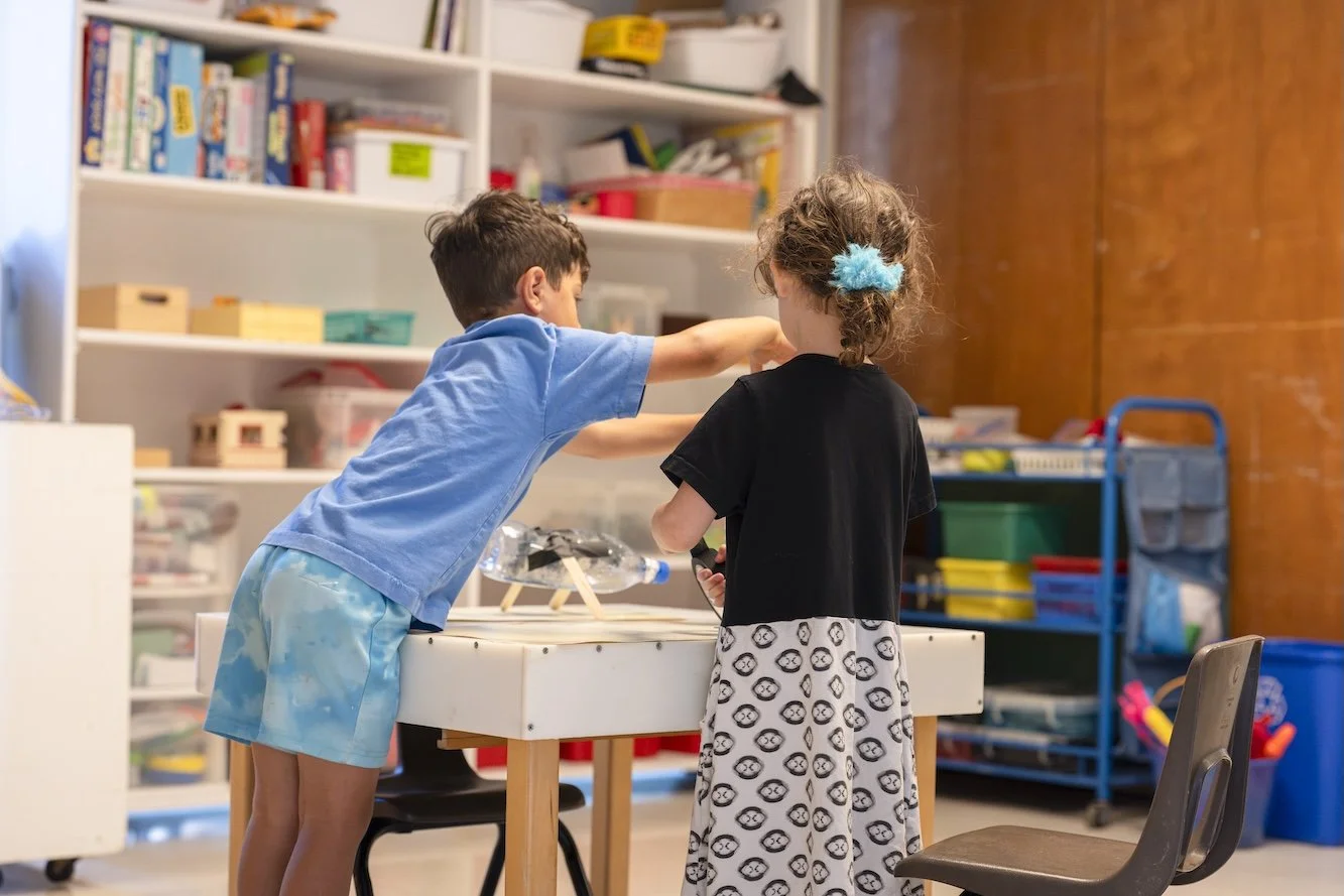 A boy and girl playing together at a table in a playroom, with shelves filled with toys and books in the background.