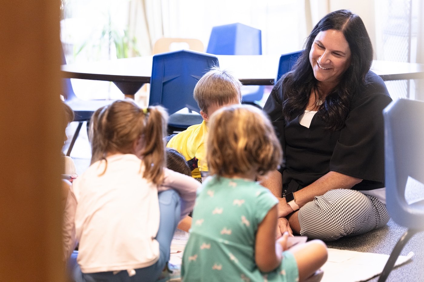 A woman sitting on the floor and smiling as she interacts with young children seated around her, in a bright room with large windows.