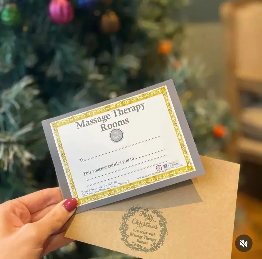 A person holding a voucher for Massage therapy rooms in front of a decorated Christmas tree. Any Gift Vouchers or E- Vouchers can be purchased on line or in store.