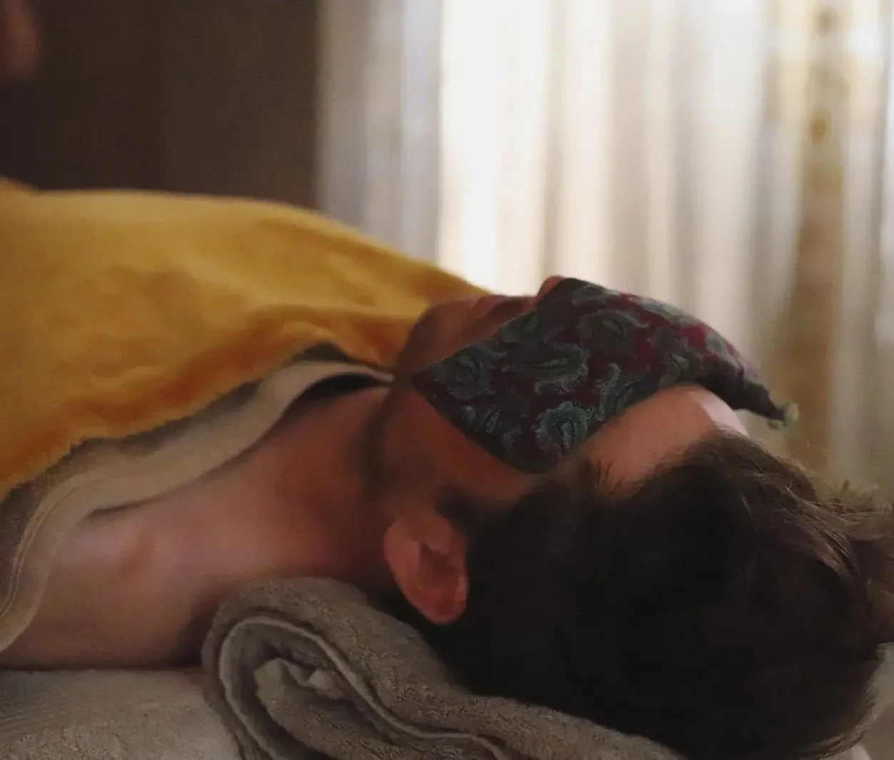 Man lying on a bed with head resting on a rolled towel, wearing a face mask and a yellow blanket, in a softly lit room. Man having a Man kind facial and Reiki treatment.