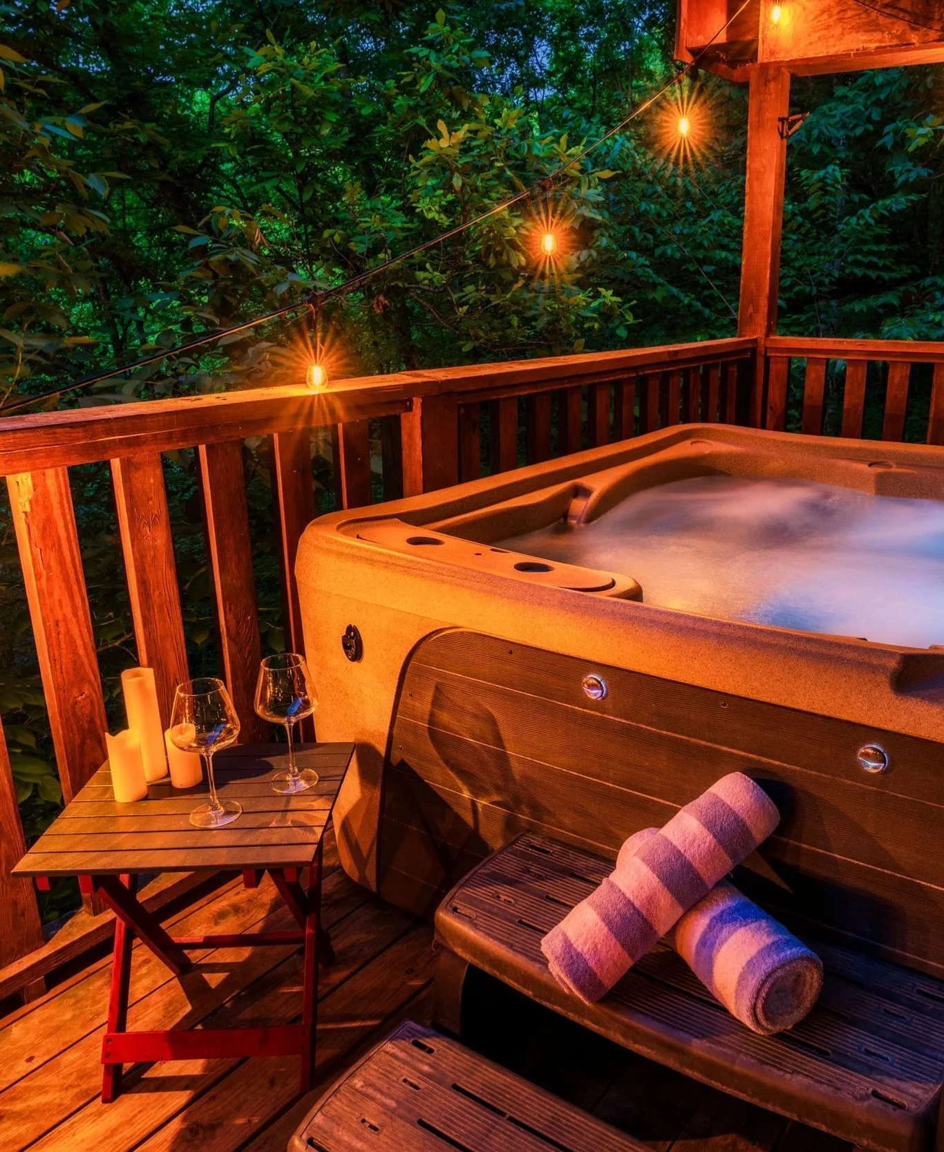 Blue Sage Hideaway's upper level deck with a hot tub, illuminated by string lights, surrounded by green trees, with a small wooden table holding two glasses of wine and candles, and a rolled towel placed on the hot tub's edge.