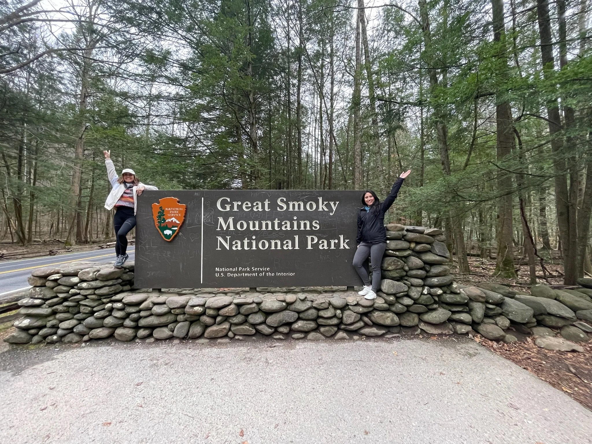 Two women standing beside a sign that reads 'Great Smoky Mountains National Park,' surrounded by a forest of tall green trees.