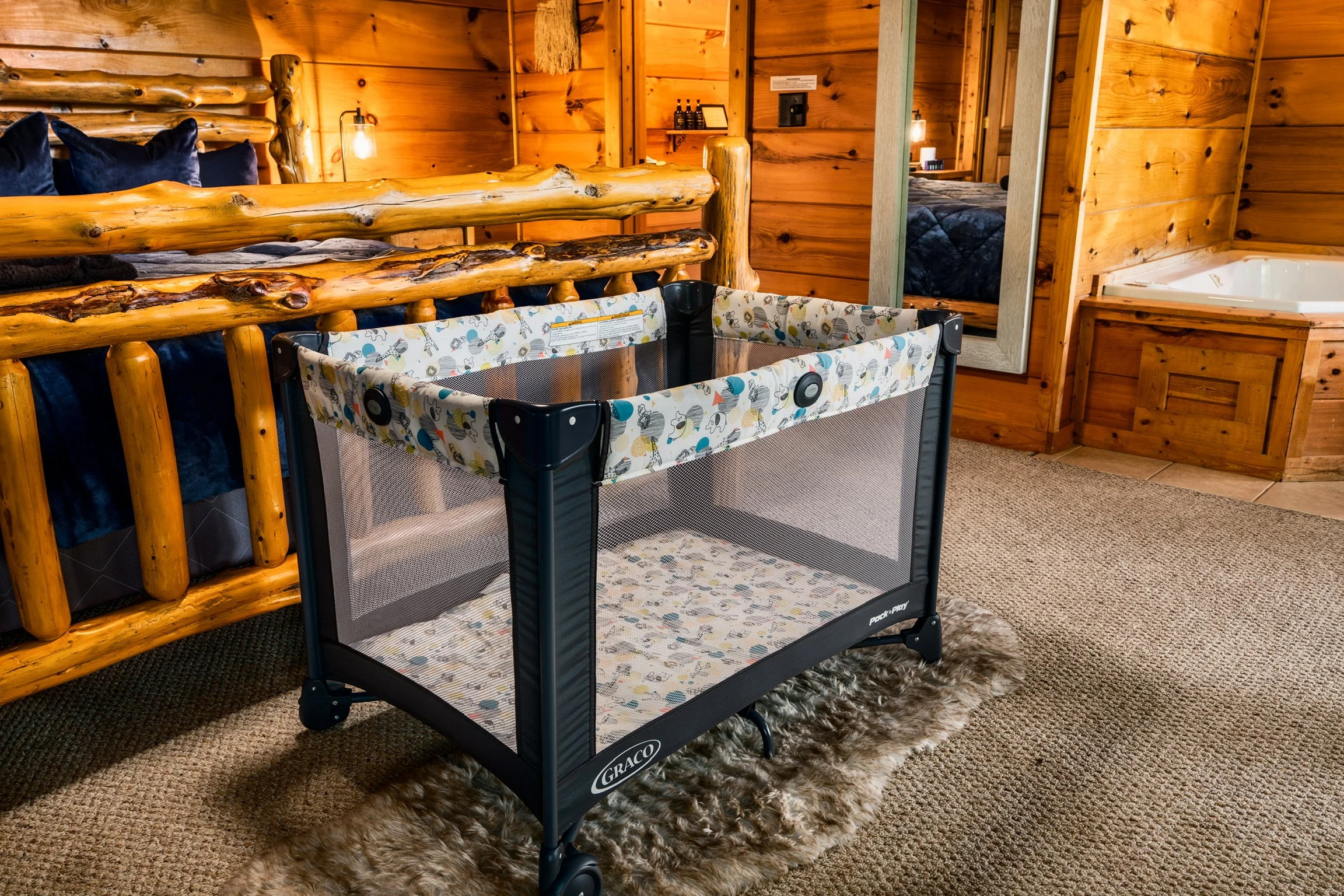 The image shows a cozy indoor space with wooden walls and furniture, featuring a wooden bed with dark blue pillows, a mirror, and a changing table or crib with playful animal print fabric. A section of a bathroom or additional room with a bathtub is visible in the background.