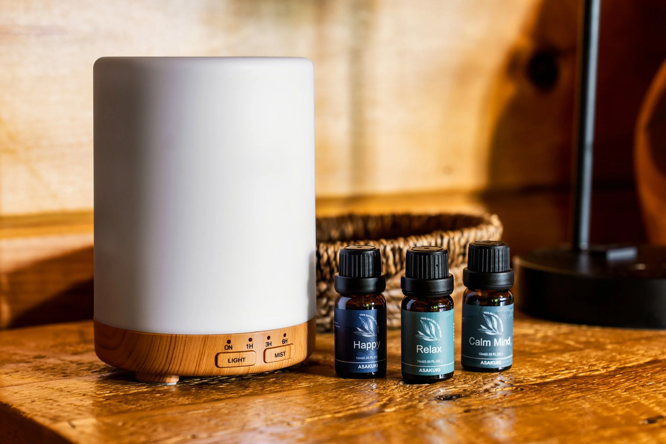 A white diffuser with a wooden base, four black essential oil bottles labeled "Happy," "Relax," "Calm Mind," and one with a partially visible label, all on a wooden surface.