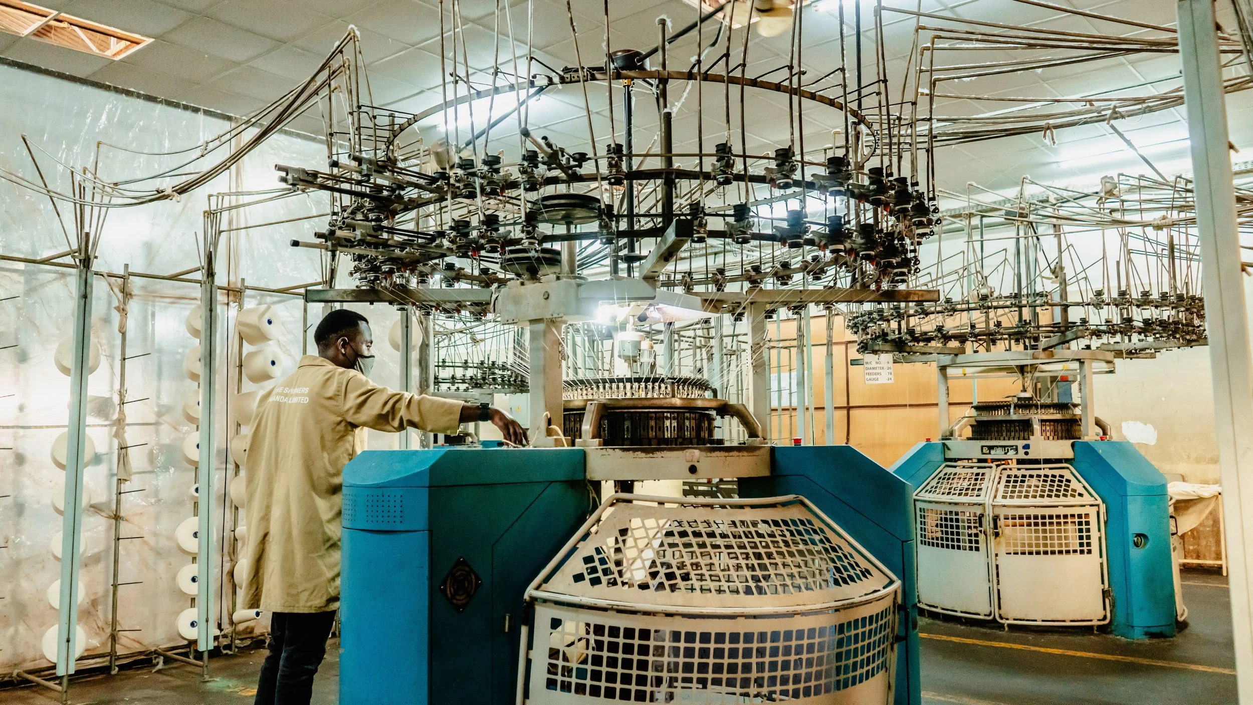 A person wearing a face mask operates industrial knitting machinery in a textile factory. The machinery includes multiple spools and metal rods used for yarn or fiber processing.