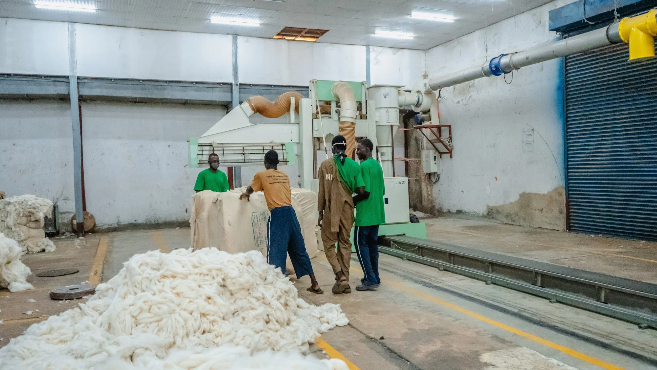 Five workers in a factory are handling raw cotton next to a large industrial machine. One worker is wearing glasses and a green shirt, while another is wearing a brown shirt. The factory has a white wall, a gray ceiling, and sunlight coming through an opening in the ceiling.