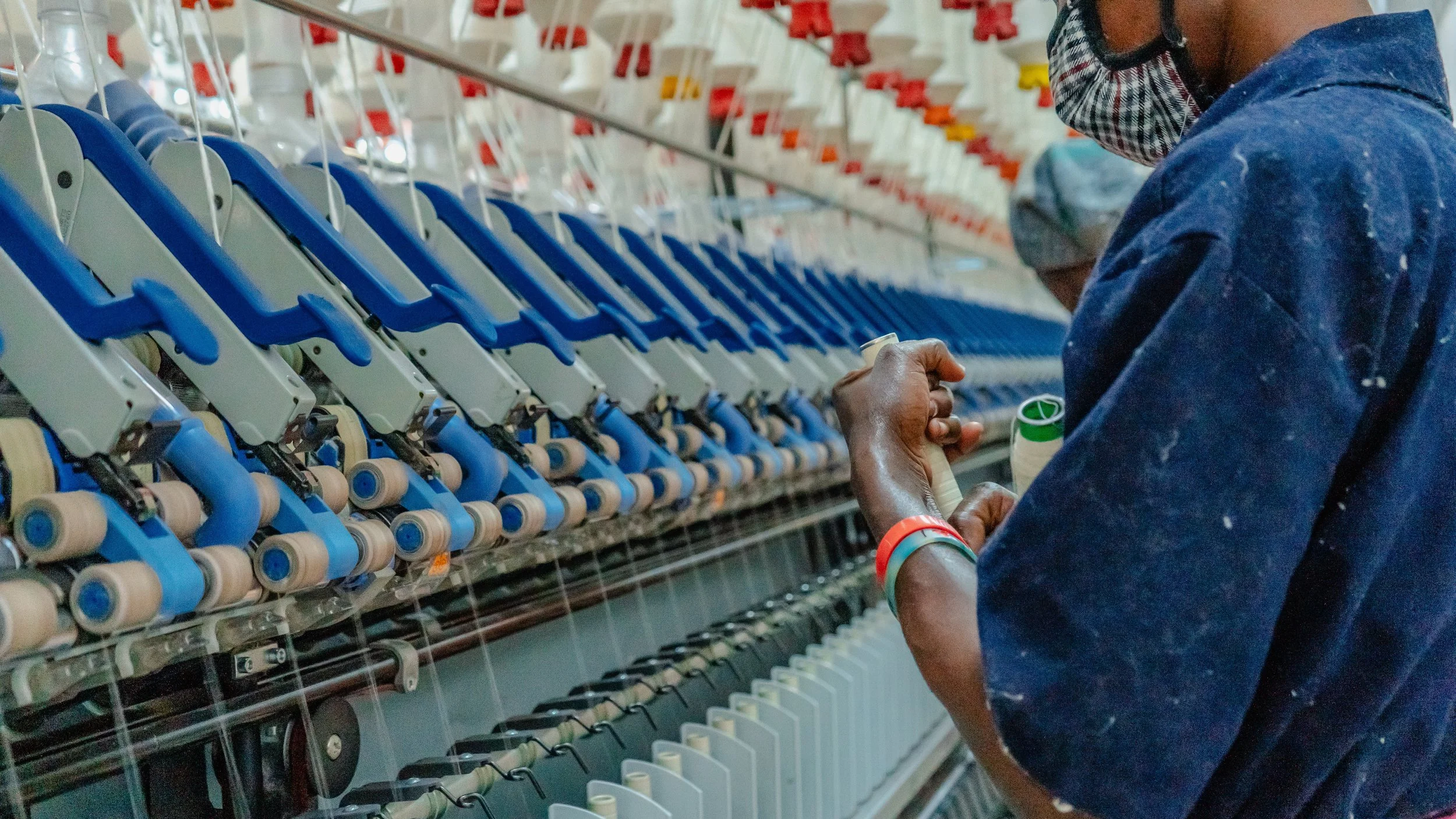 A person with dark skin, wearing a face mask, a blue shirt, and colorful bracelets, is working on a textile machine with spools of thread, in a factory setting.