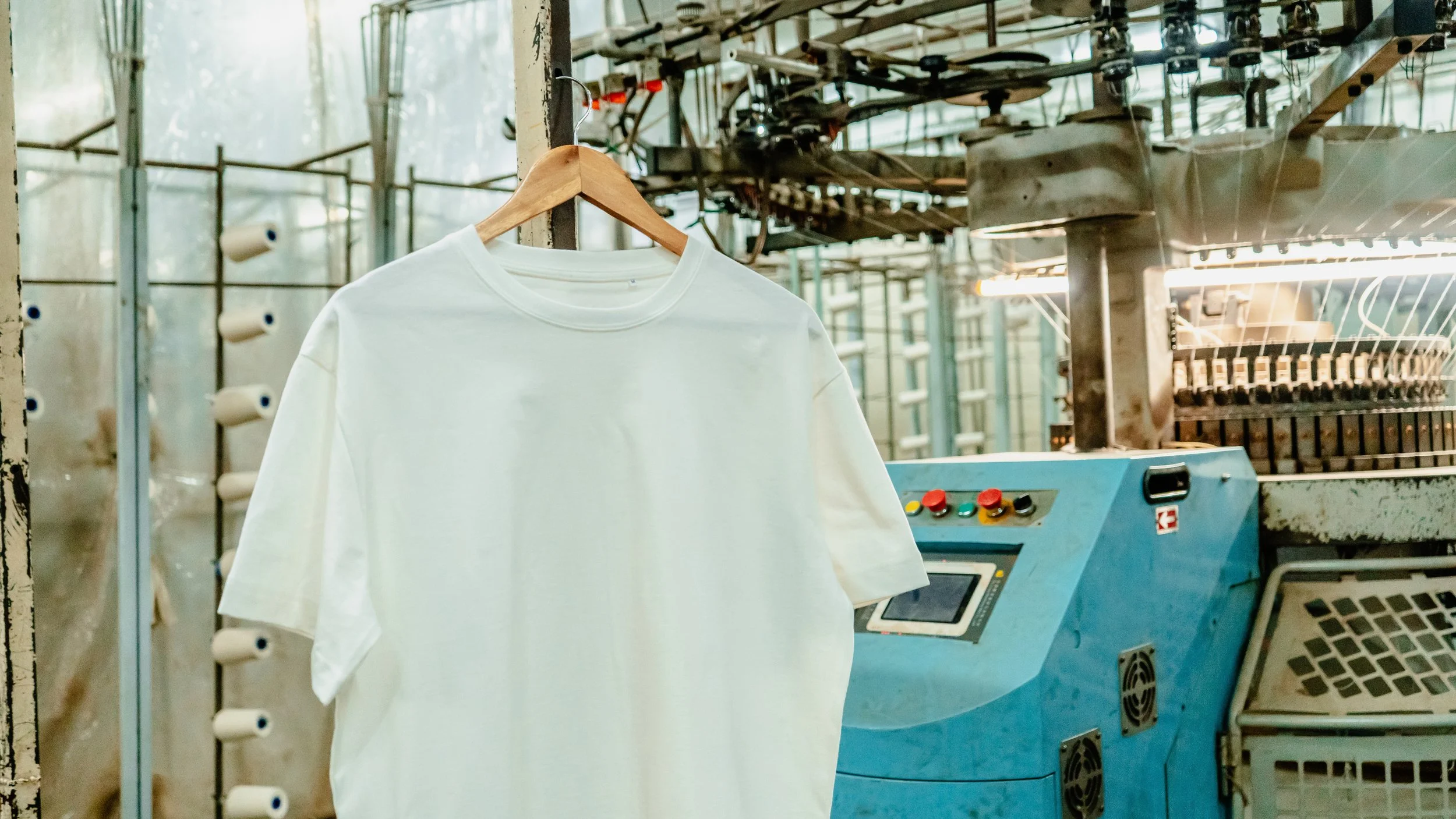 A white T-shirt hanging on a wooden hanger in front of textile machinery in a factory.