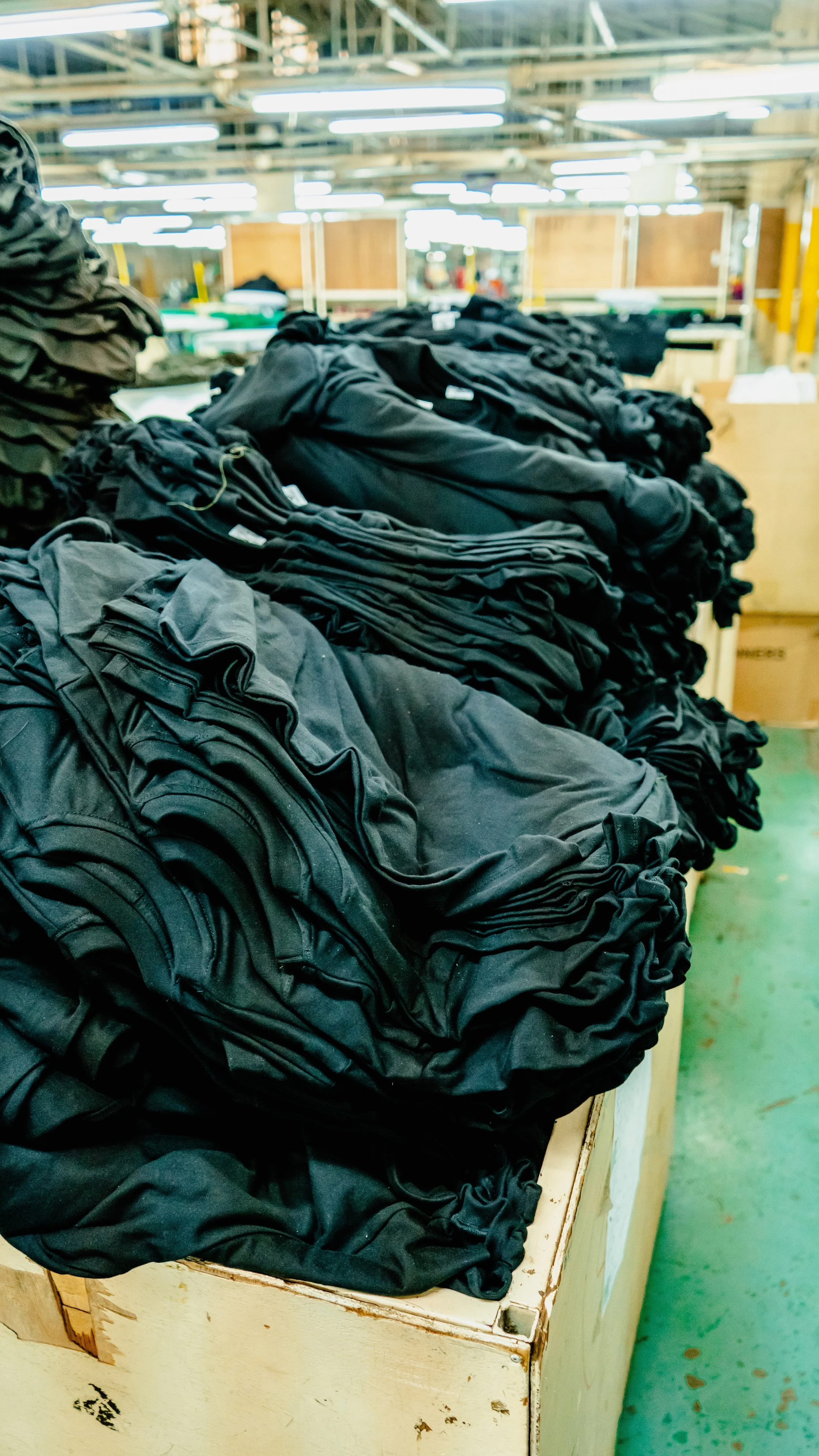 Stacks of black and gray clothing, possibly T-shirts or hoodies, on a wooden table in what appears to be a factory or warehouse.
