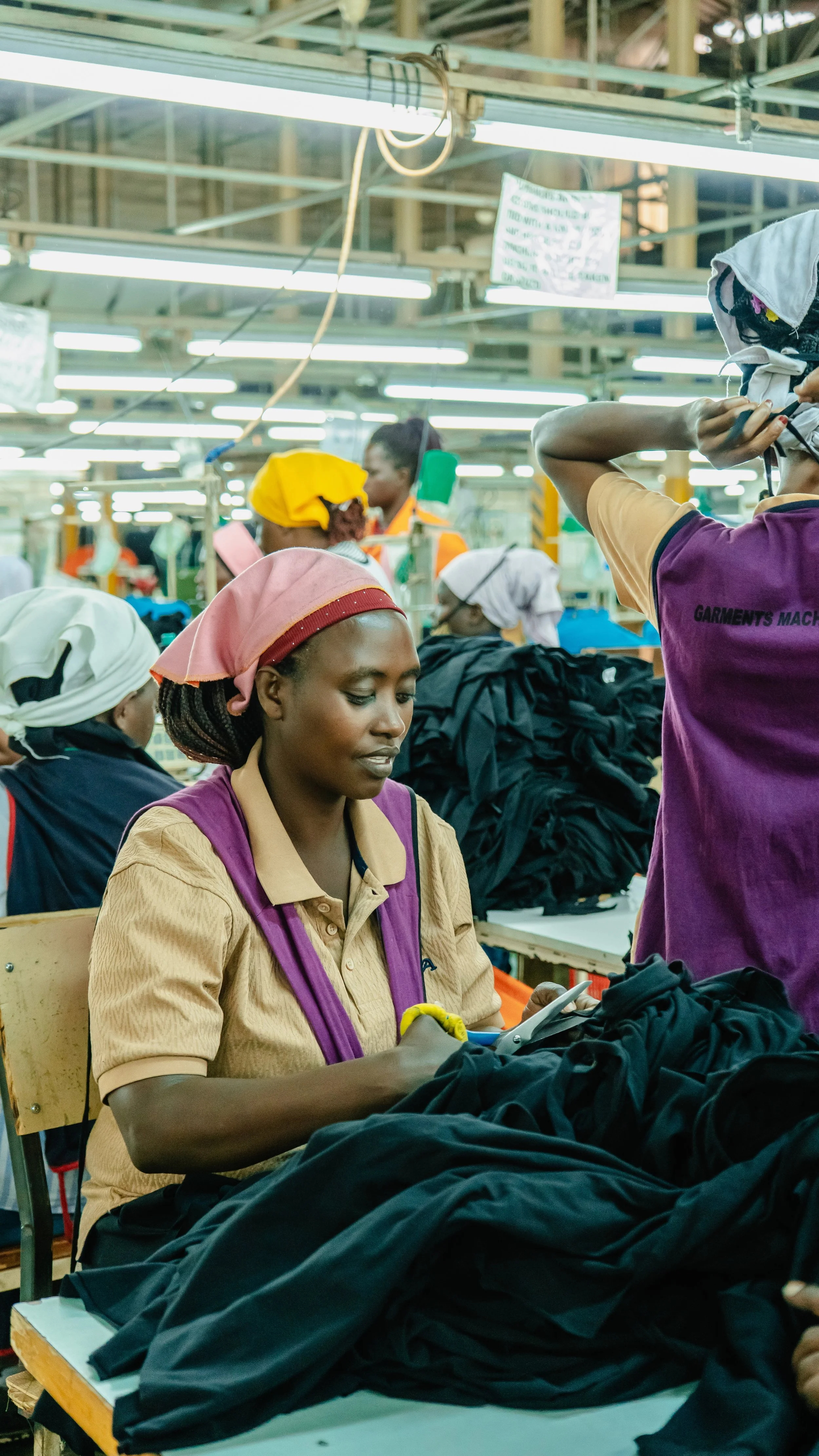 Workers in a garment factory sewing black fabric with sewing machines, surrounded by piles of fabric and workers wearing head coverings.