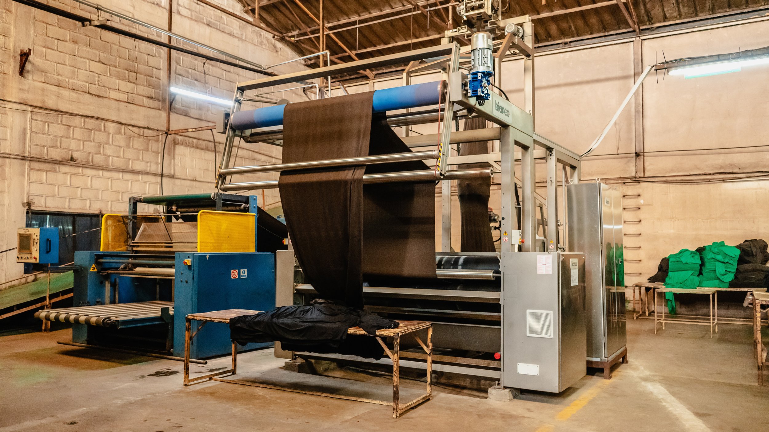 Industrial textile manufacturing machine in a warehouse with stacks of fabric and green clothing to the side.