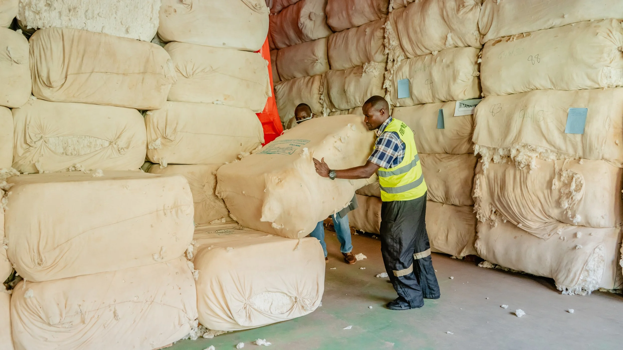 Workers handling large bales of cotton in an industrial storage area.