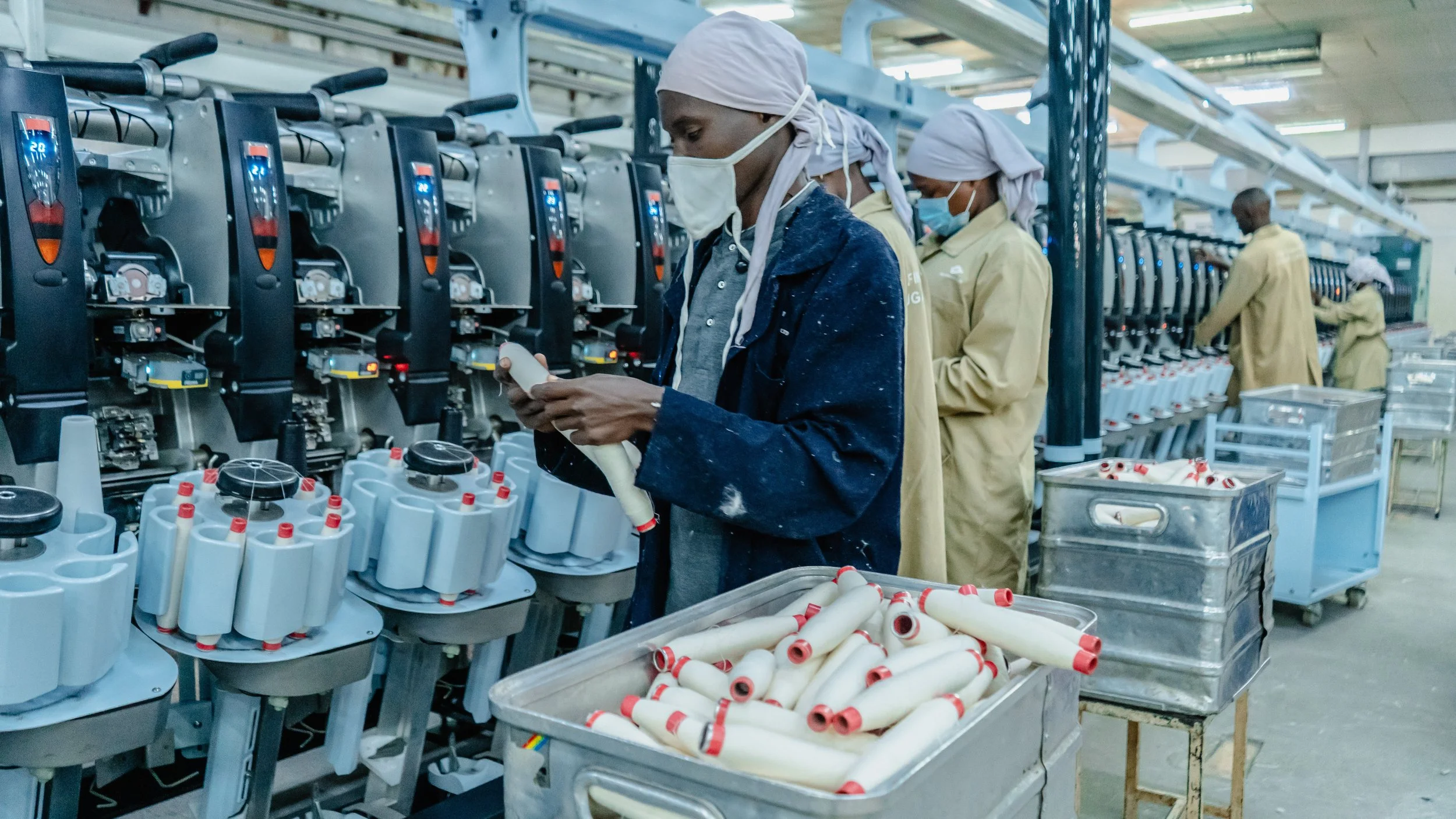 Workers in a factory wearing masks and hair coverings, assembling or inspecting electronic or mechanical components on production lines.