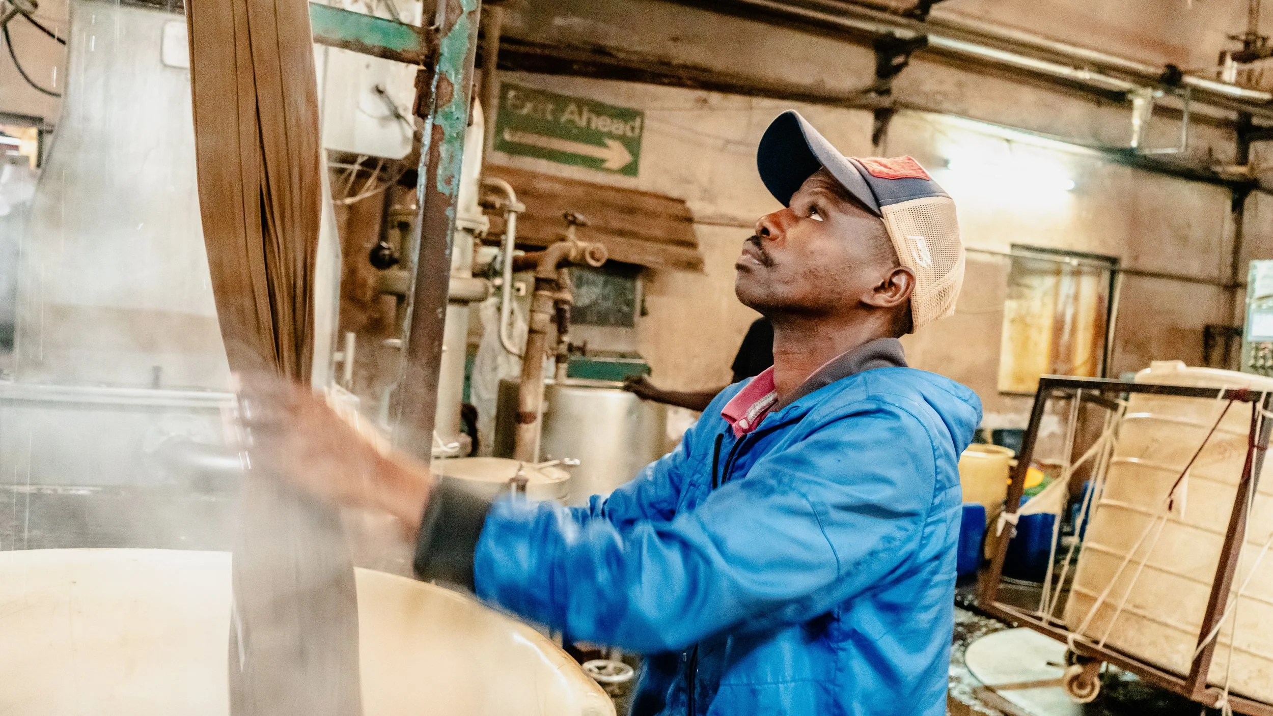 A man in a blue jacket and baseball cap working in a rustic workshop surrounded by tools and wooden materials, with a green sign pointing right that says 'Eat Ahead' in the background.