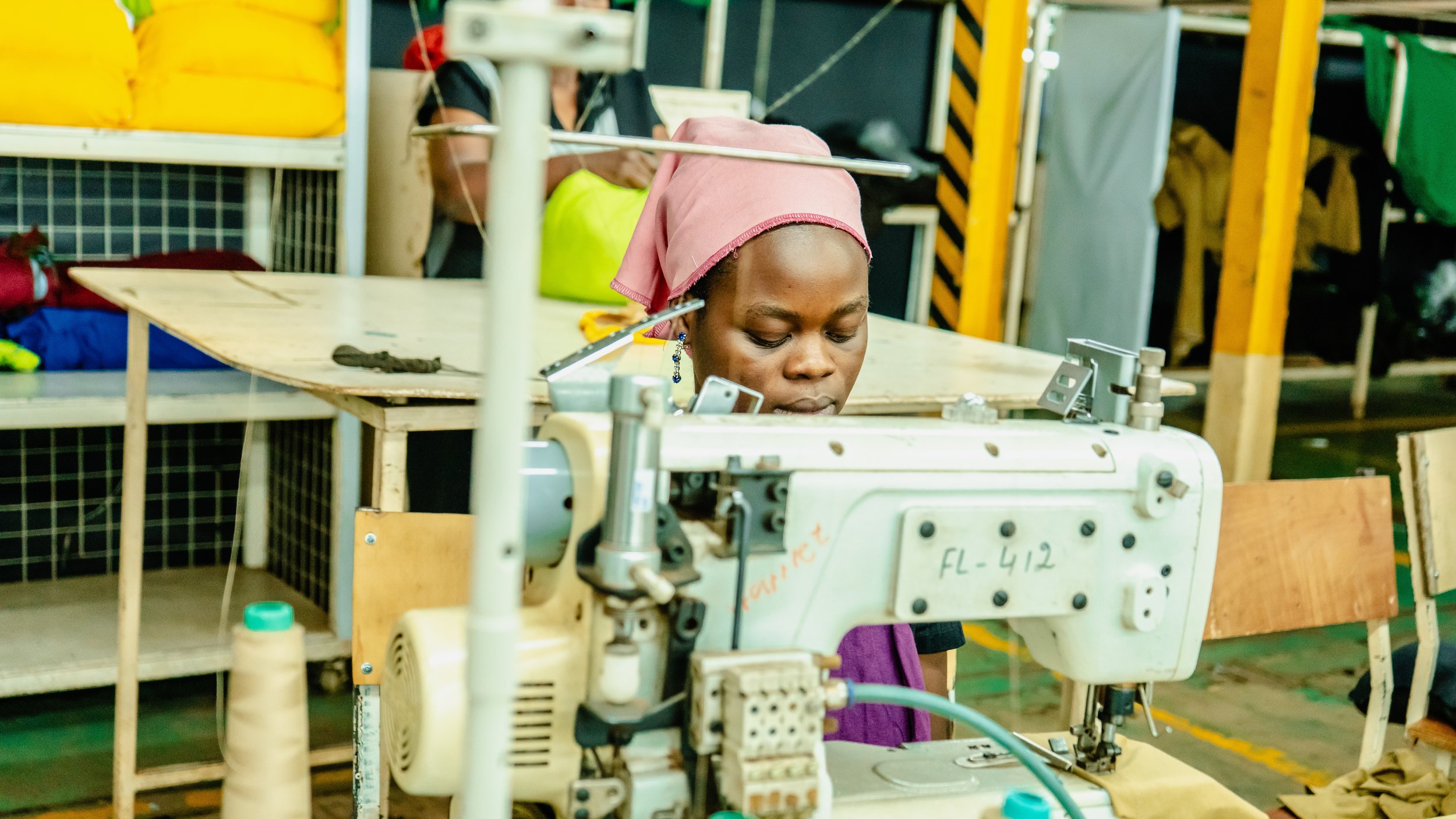 Woman working with a sewing machine in a factory or workshop, wearing a pink headscarf.