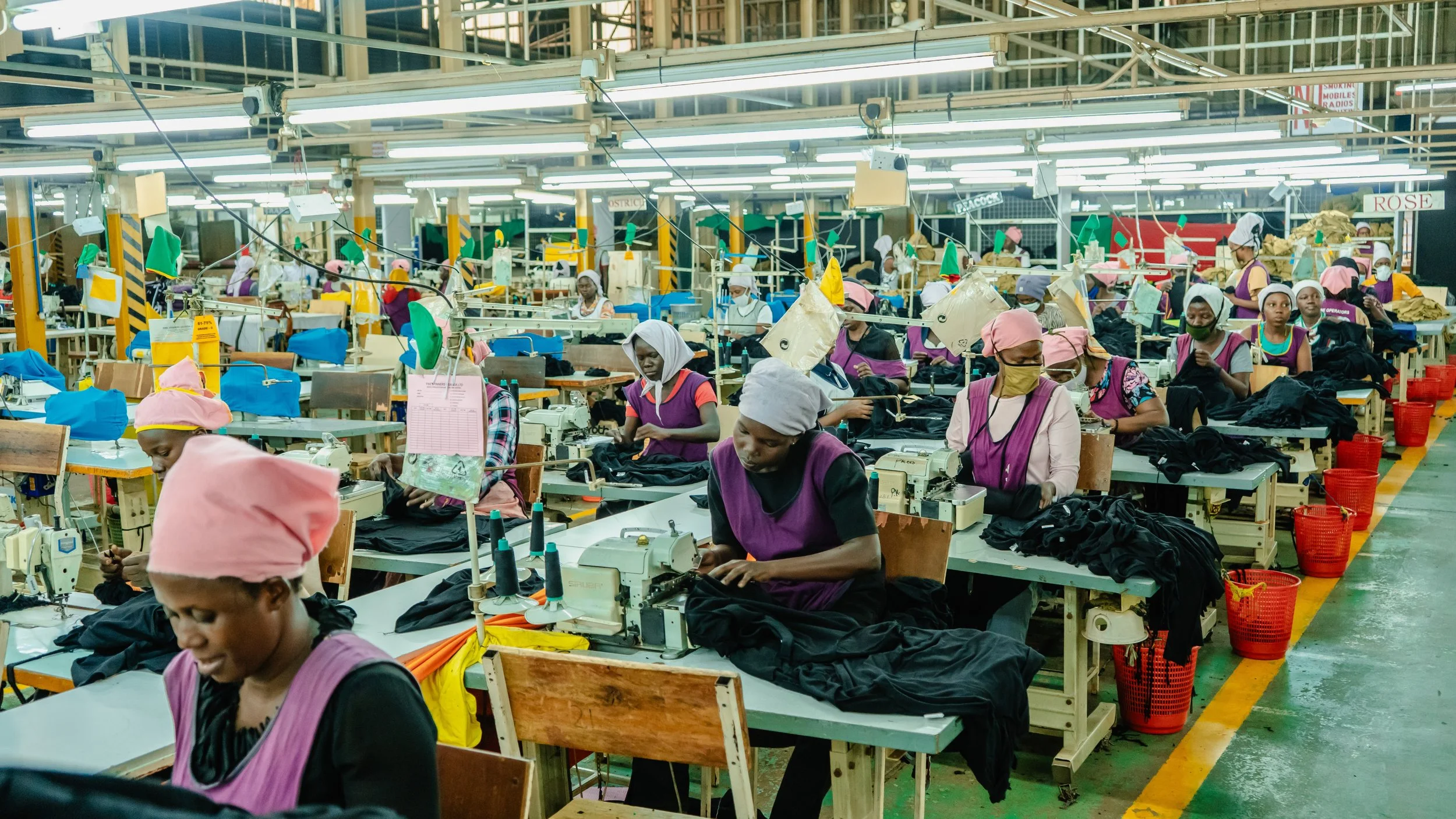 Workers in a garment factory sewing black fabric at assembly lines, wearing head coverings and masks, with bright fluorescent lights overhead.