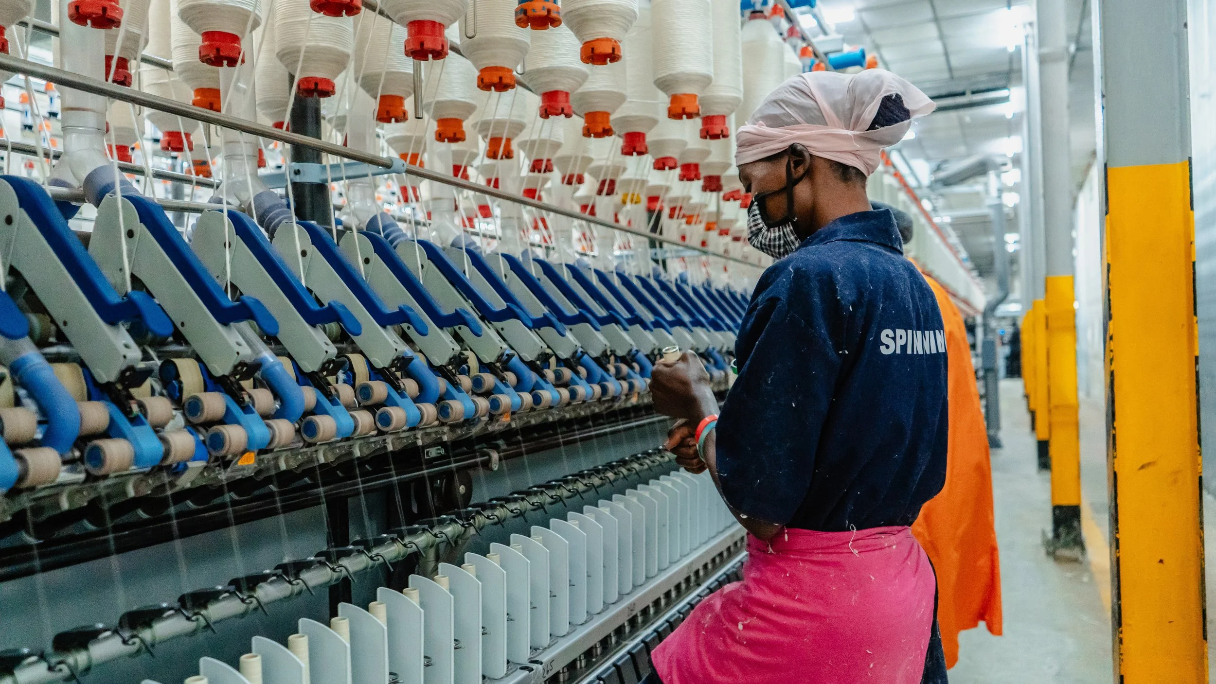 A worker in a manufacturing or textile factory is operating a machine that appears to be spinning or weaving yarn or thread. The worker is wearing a white head wrap, glasses, a face mask, a dark blue jacket with the word "SPINNING" on the back, and pink pants.