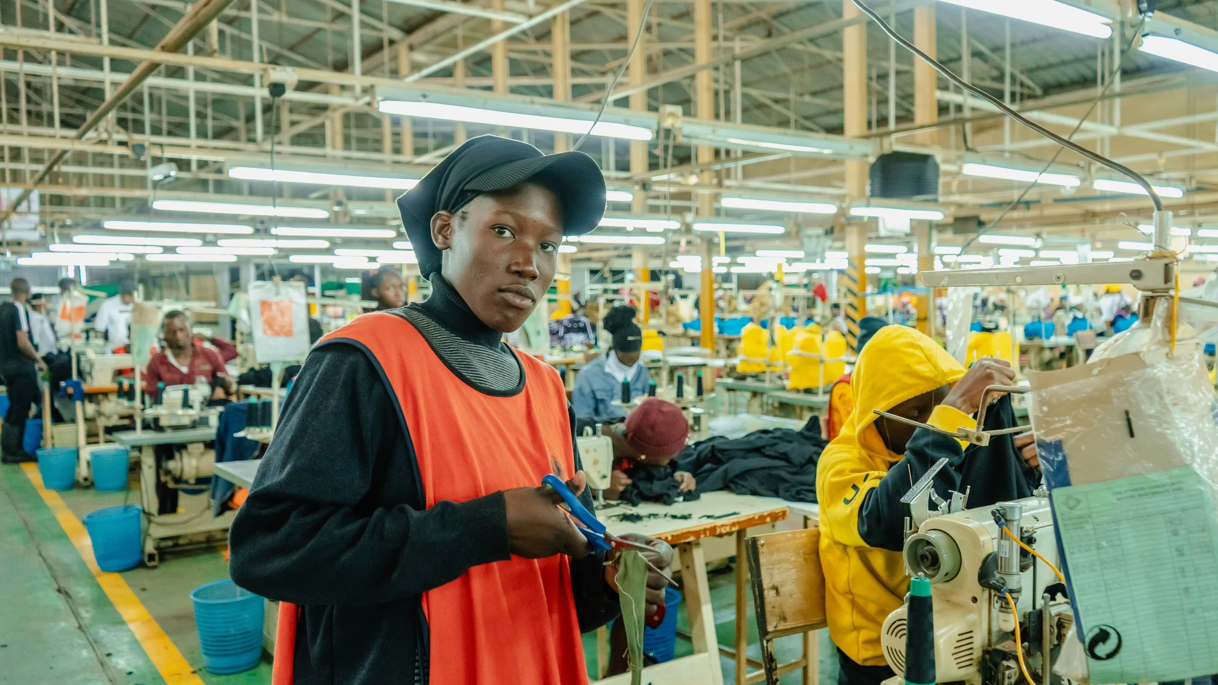 Young woman working in a factory or workshop with sewing machines, wearing a black hat and orange safety vest, surrounded by other workers and machinery.