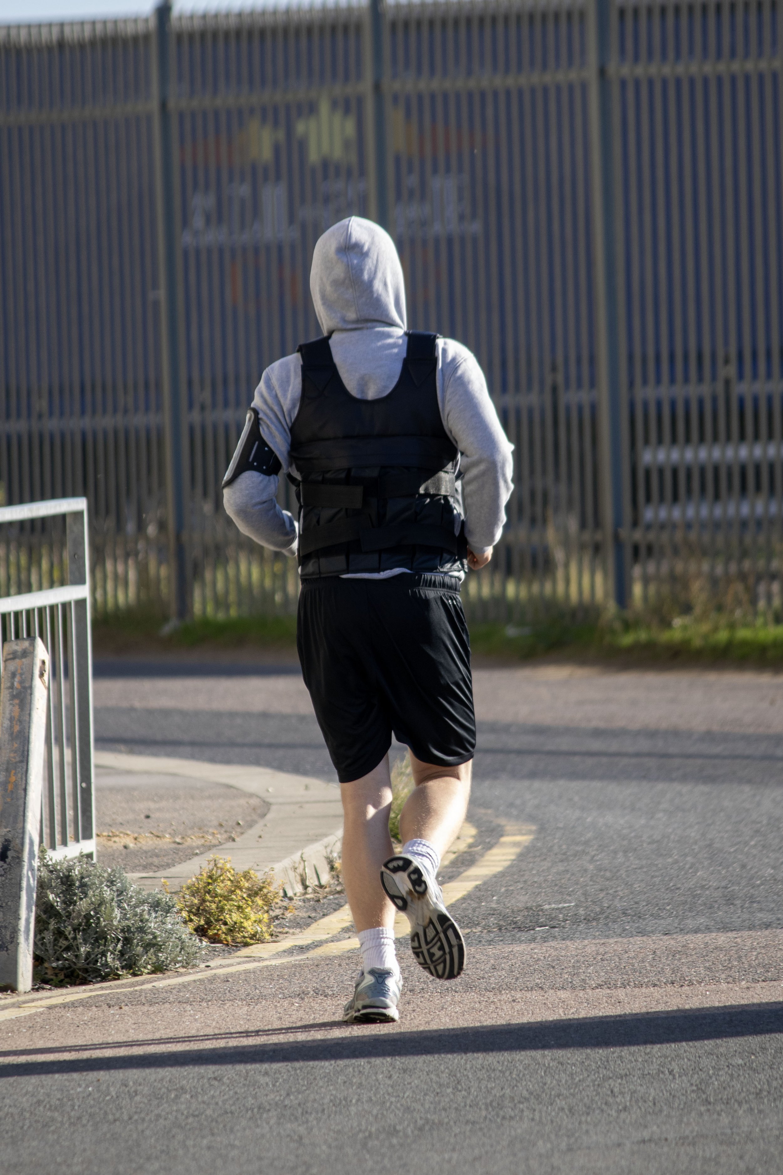 A person jogging on a sidewalk wearing a gray hoodie, black shorts, a black vest, and white sneakers, with foliage and a metal fence in the background.