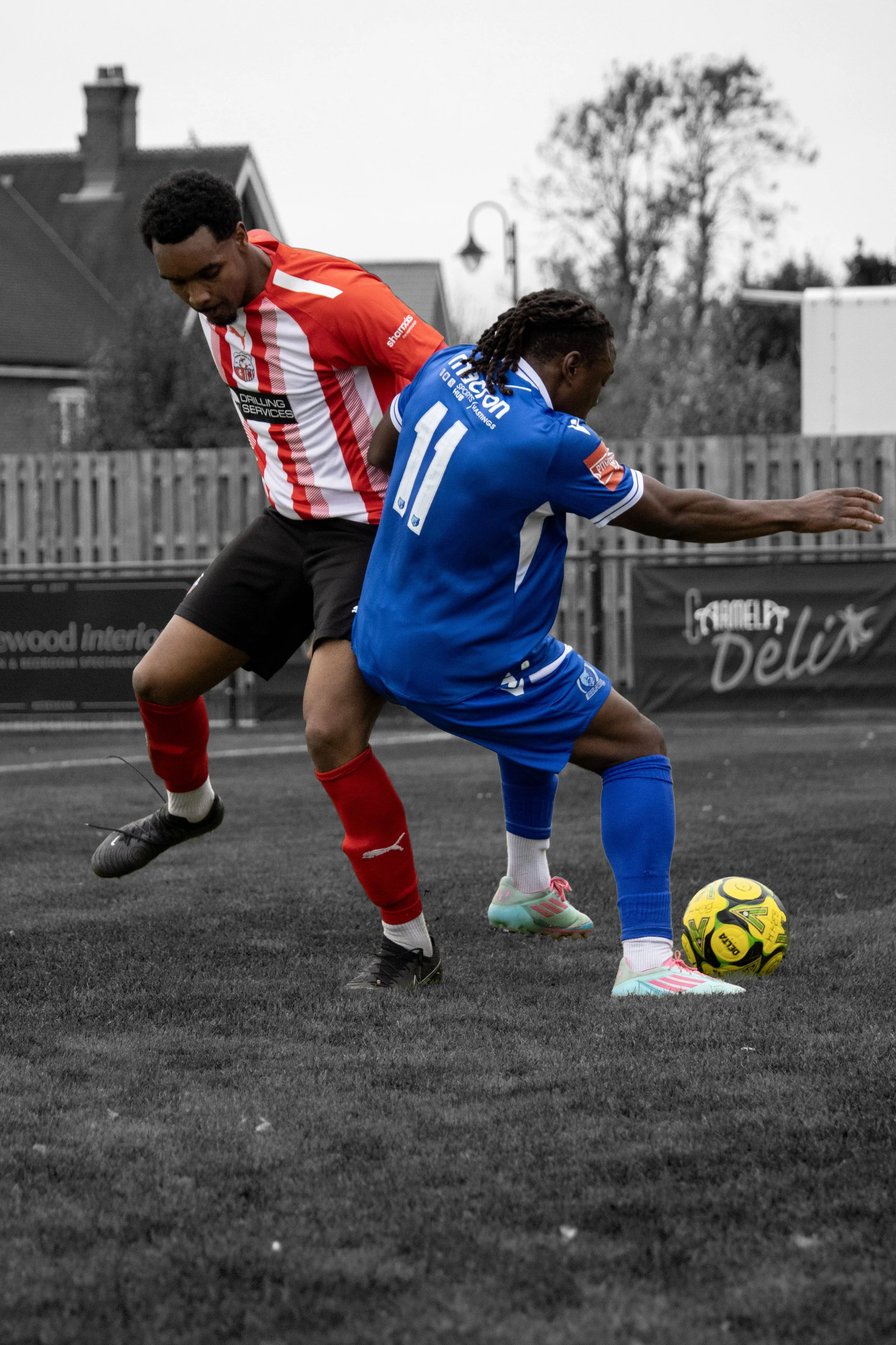 Two soccer players are competing for the ball on a soccer field, both wearing different colored jerseys and shorts, one in red and black and the other in blue and white, with a background of residential buildings and a fence.