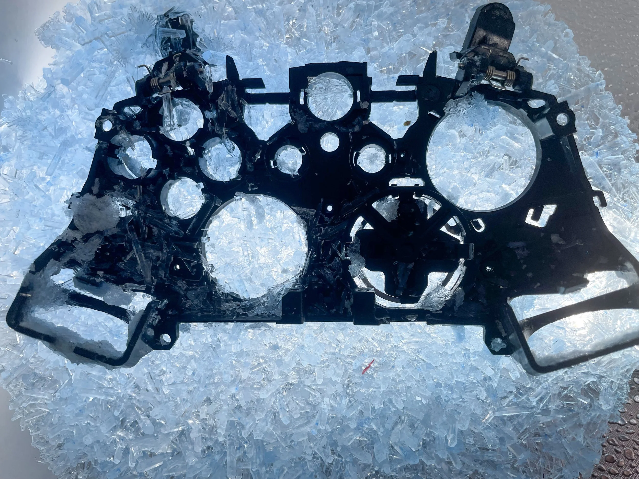 Broken glass shards arranged in a semi-circle around a black mechanical part on a reflective surface.