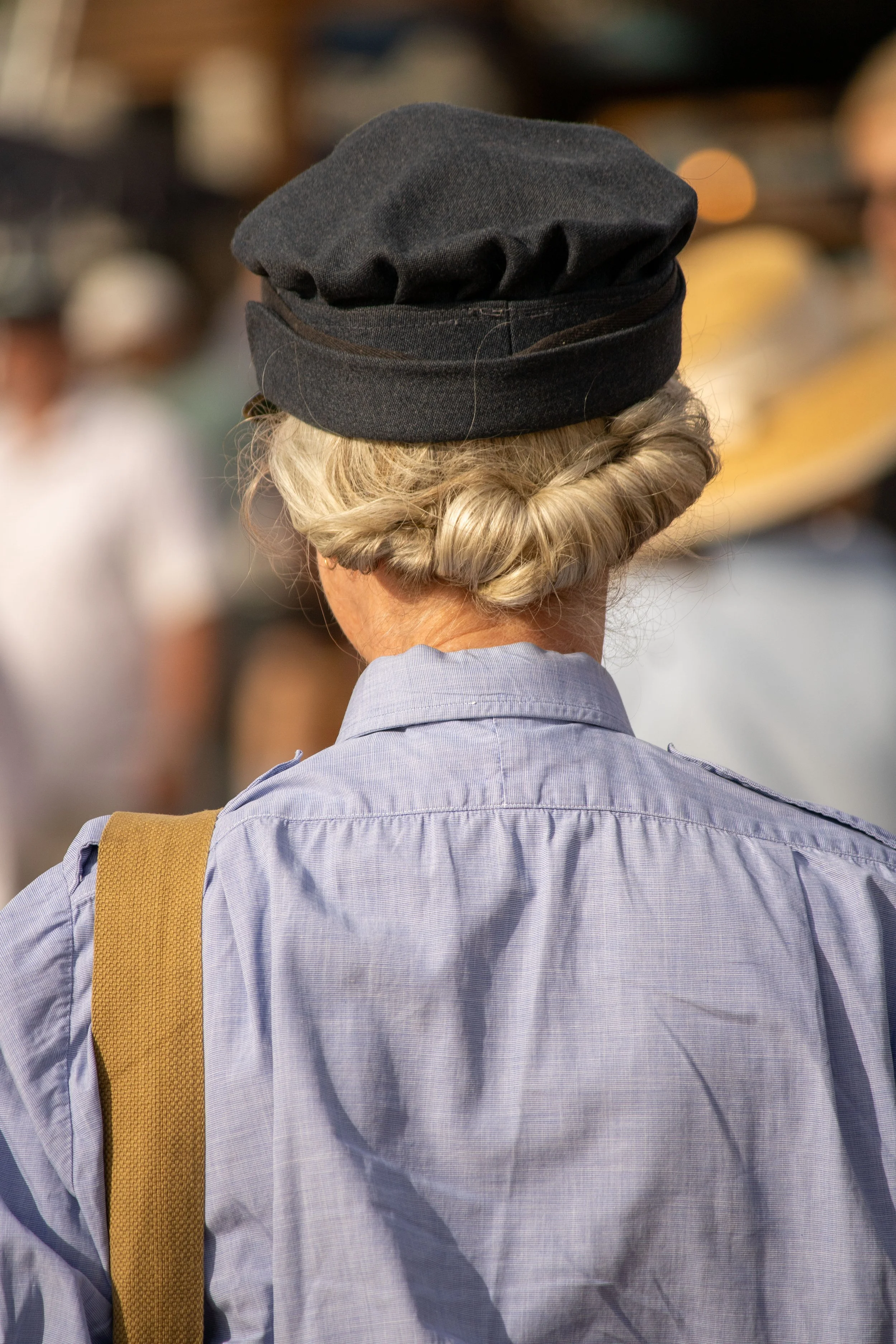 Back view of a woman with gray hair styled in a bun, wearing a black hat, a light blue button-up shirt, and a mustard-colored strap over her shoulder, standing in a blurred busy outdoor setting.