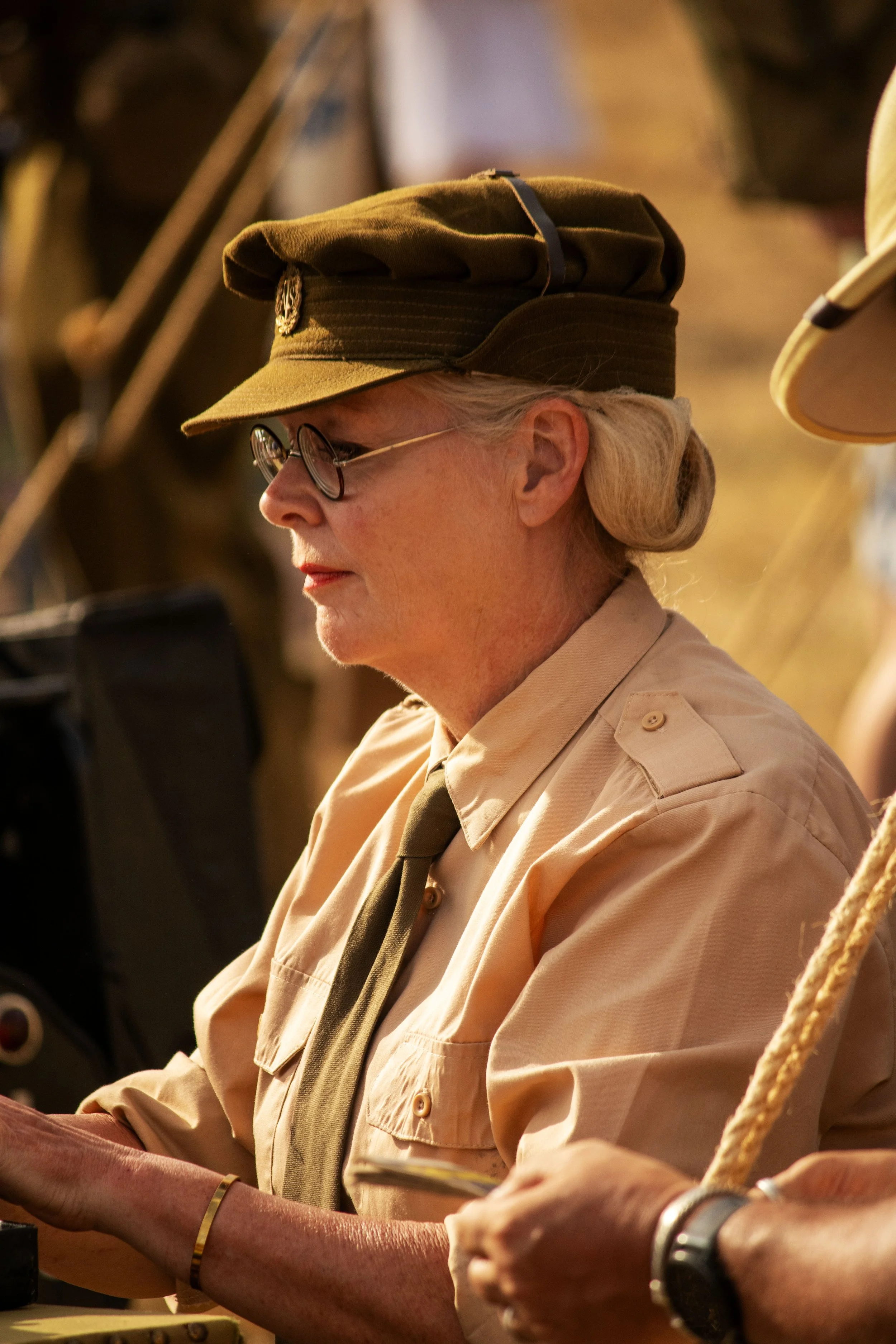 A woman with glasses and gray hair, dressed in a khaki uniform, wearing a military-style cap, sitting with her hands on a table or equipment, in a setting that appears to be outdoors or in a rustic environment.