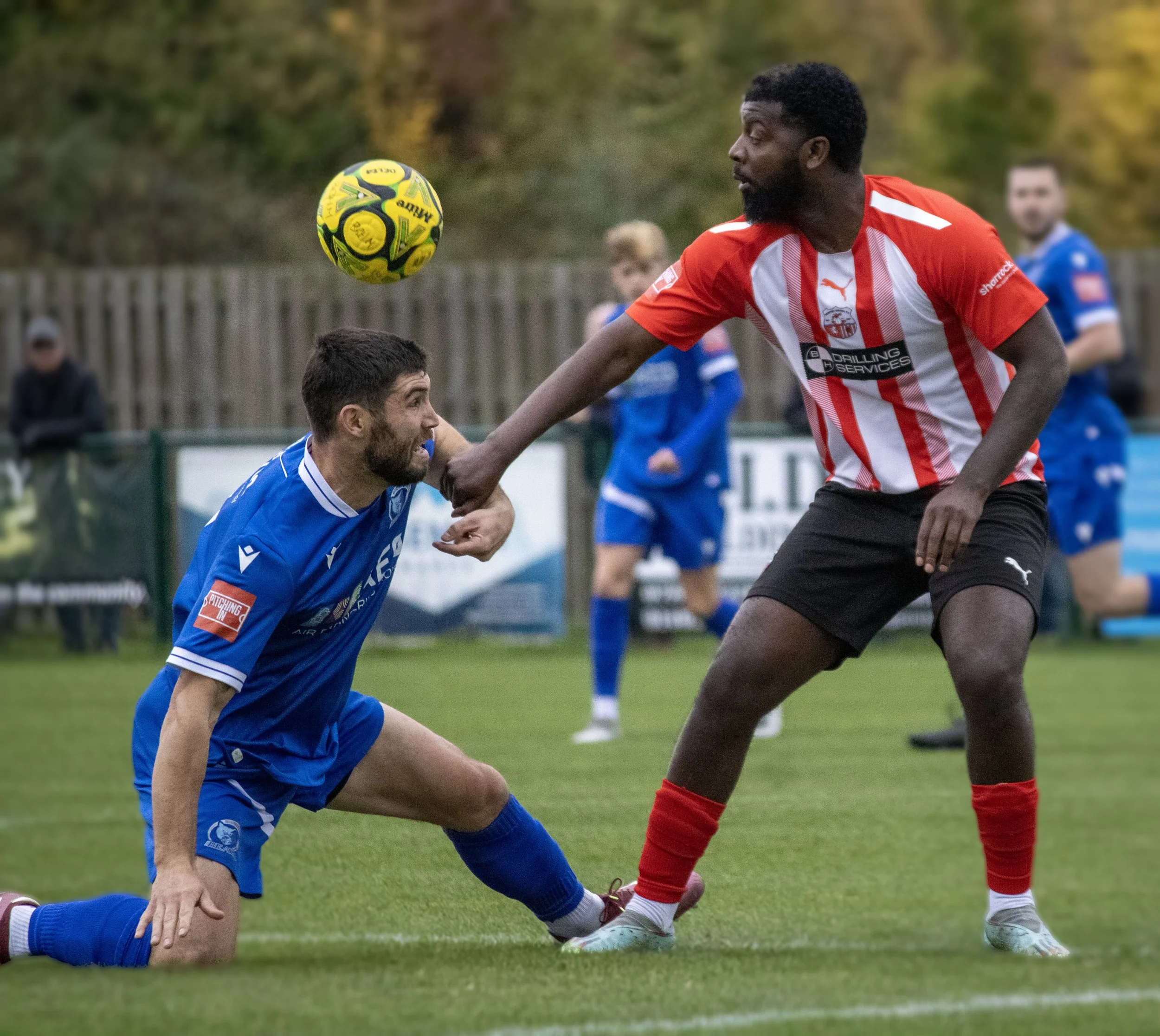 A soccer player in a red and white striped jersey and red socks is jumping to head the yellow and black soccer ball while another player in a blue jersey and blue shorts is on the ground, looking upwards. The scene is outdoors on a grassy field with a blurred background and a few people watching.