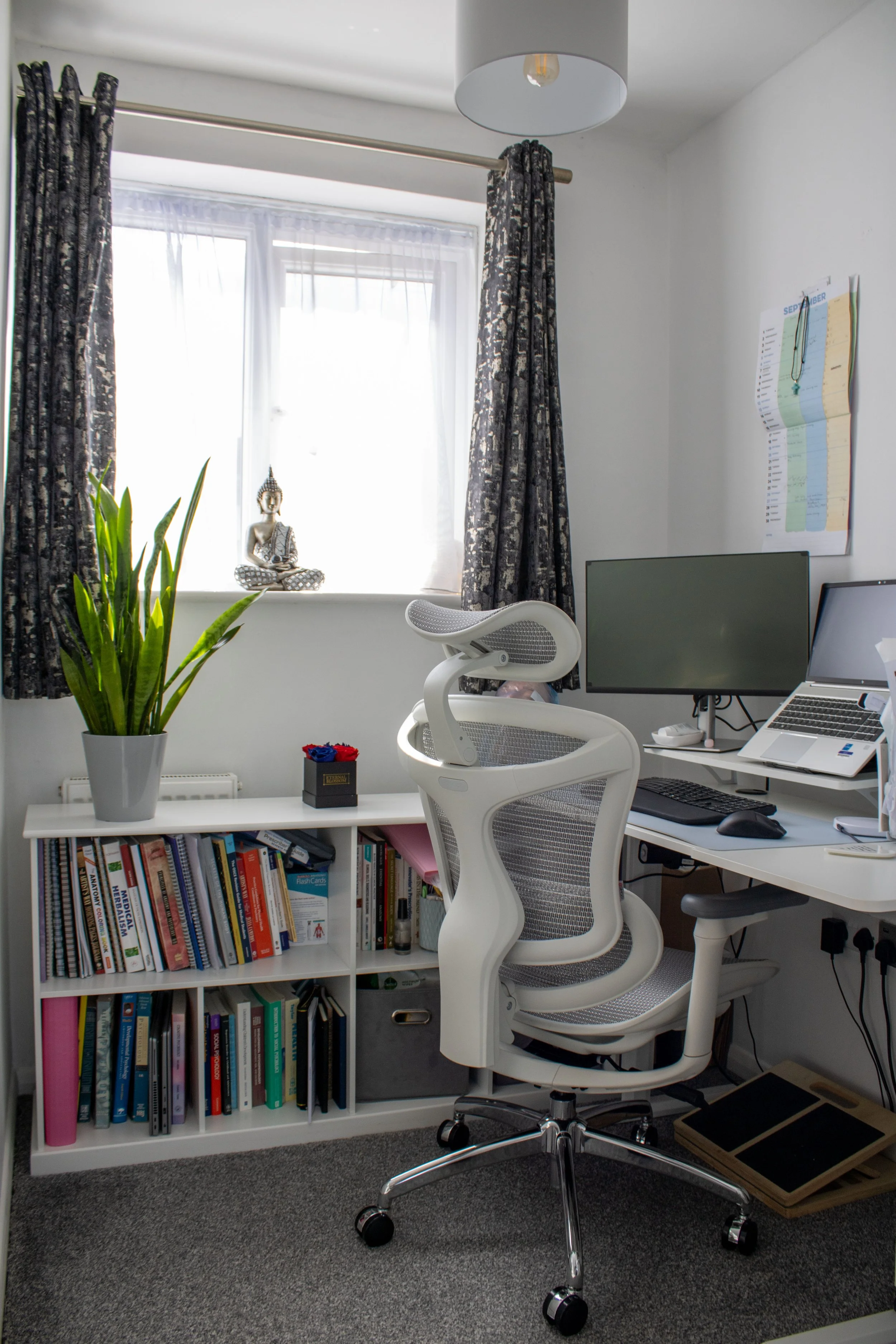 An organized home office with a white desk, an ergonomic white office chair, a large monitor, a laptop, and a bookshelf filled with colorful books. A potted snake plant and a small Buddha statue are on the window sill, accompanied by black curtains with a metallic pattern and sheer white curtains. There is a pendant light, a calendar on the wall, and a foot pedal on the floor.