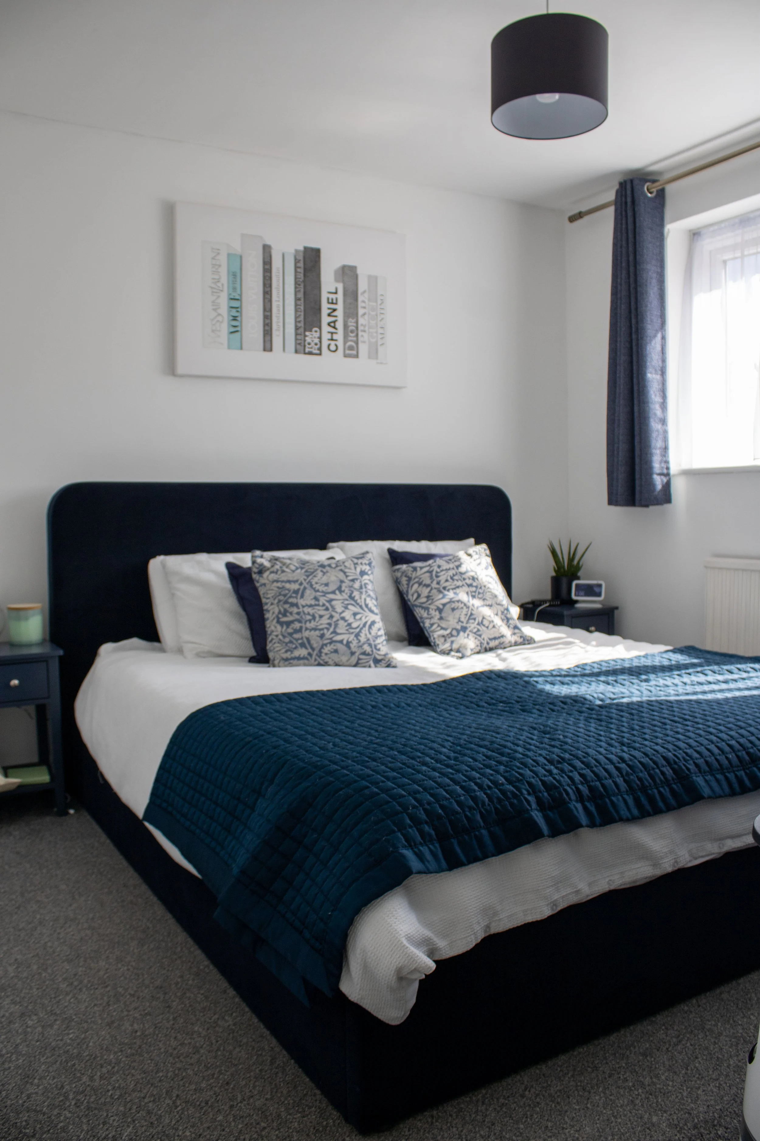 A bedroom with a bed that has a navy blue quilted blanket and decorative pillows, a dark blue headboard, a window with blue curtains, and a white wall art piece with books and brand names like Chanel and Dior.
