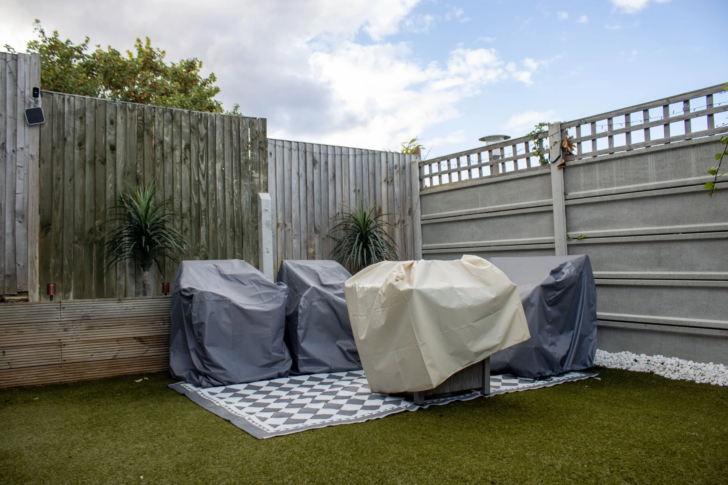 Backyard patio area with covered furniture, two palm plants, a patterned outdoor rug, wooden privacy fence, and a partly cloudy sky.