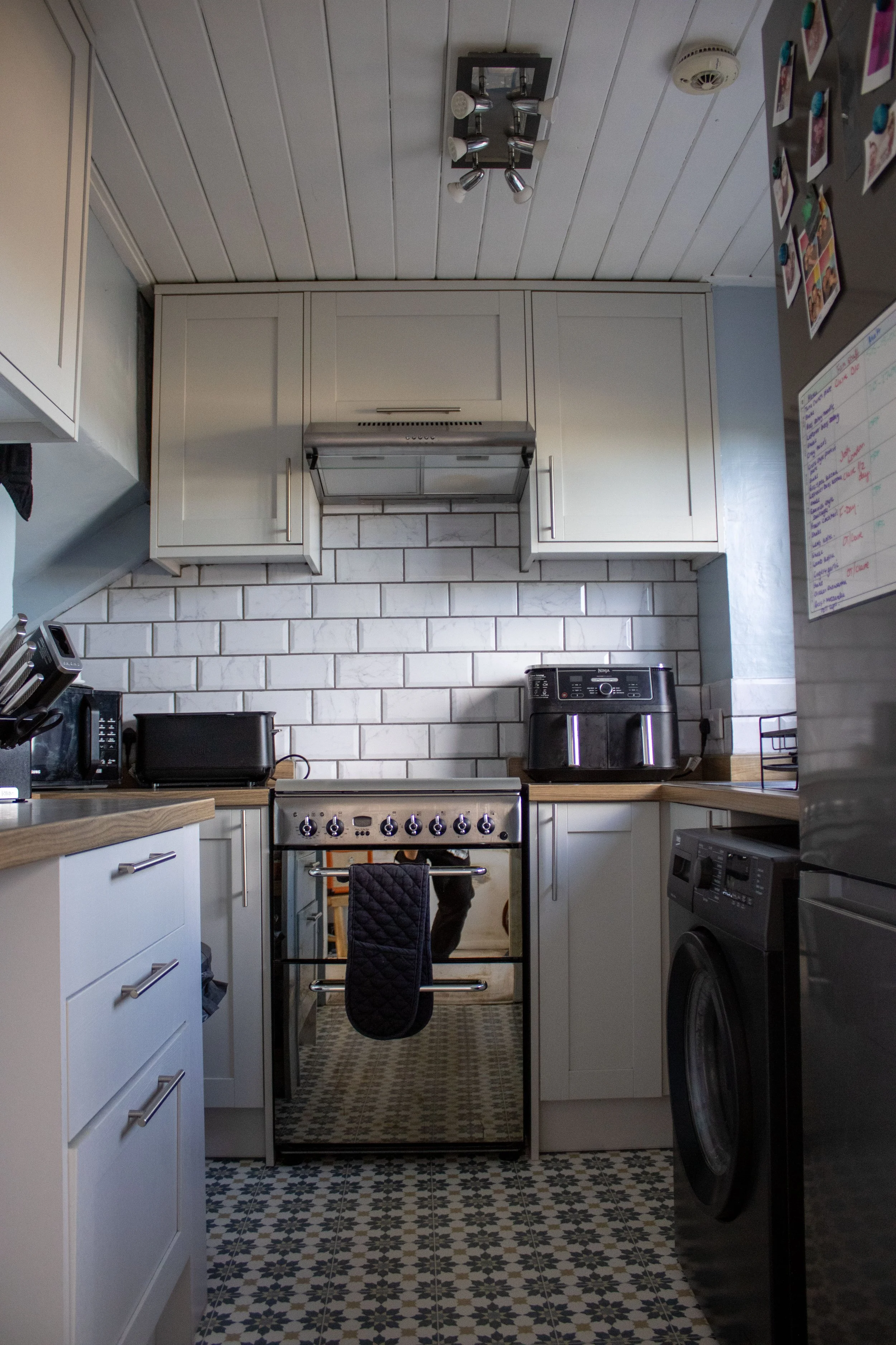 A view of a kitchen with white cabinets, a stove in the center, and a refrigerator on the right. The ceiling has wooden panels, a modern ceiling light fixture, and a smoke detector. The kitchen has a patterned tile floor and a white brick backsplash.