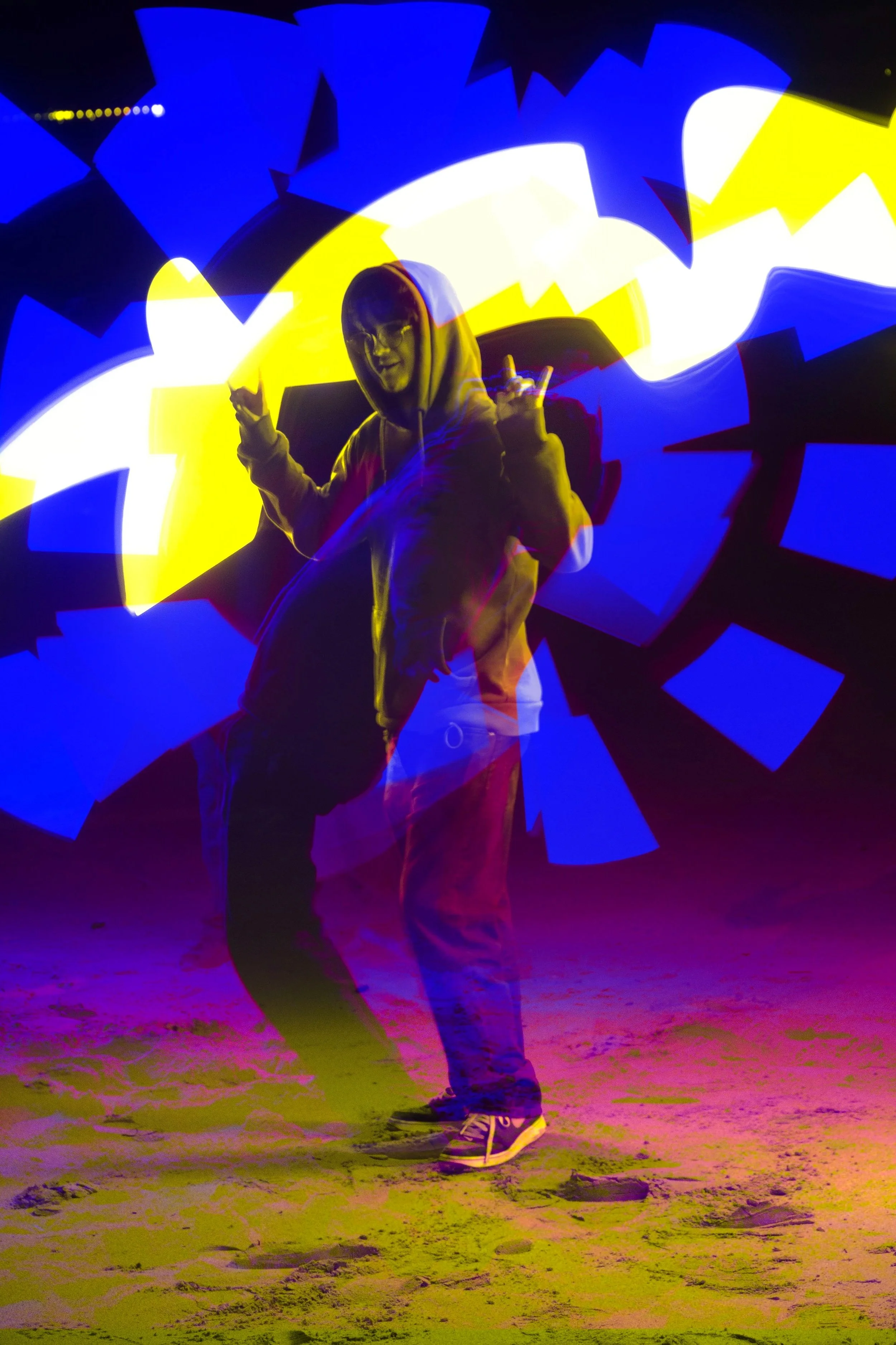 A person wearing a hoodie and sneakers standing on a sandy beach at night, surrounded by bright, colorful light patterns in blue, yellow, and purple.