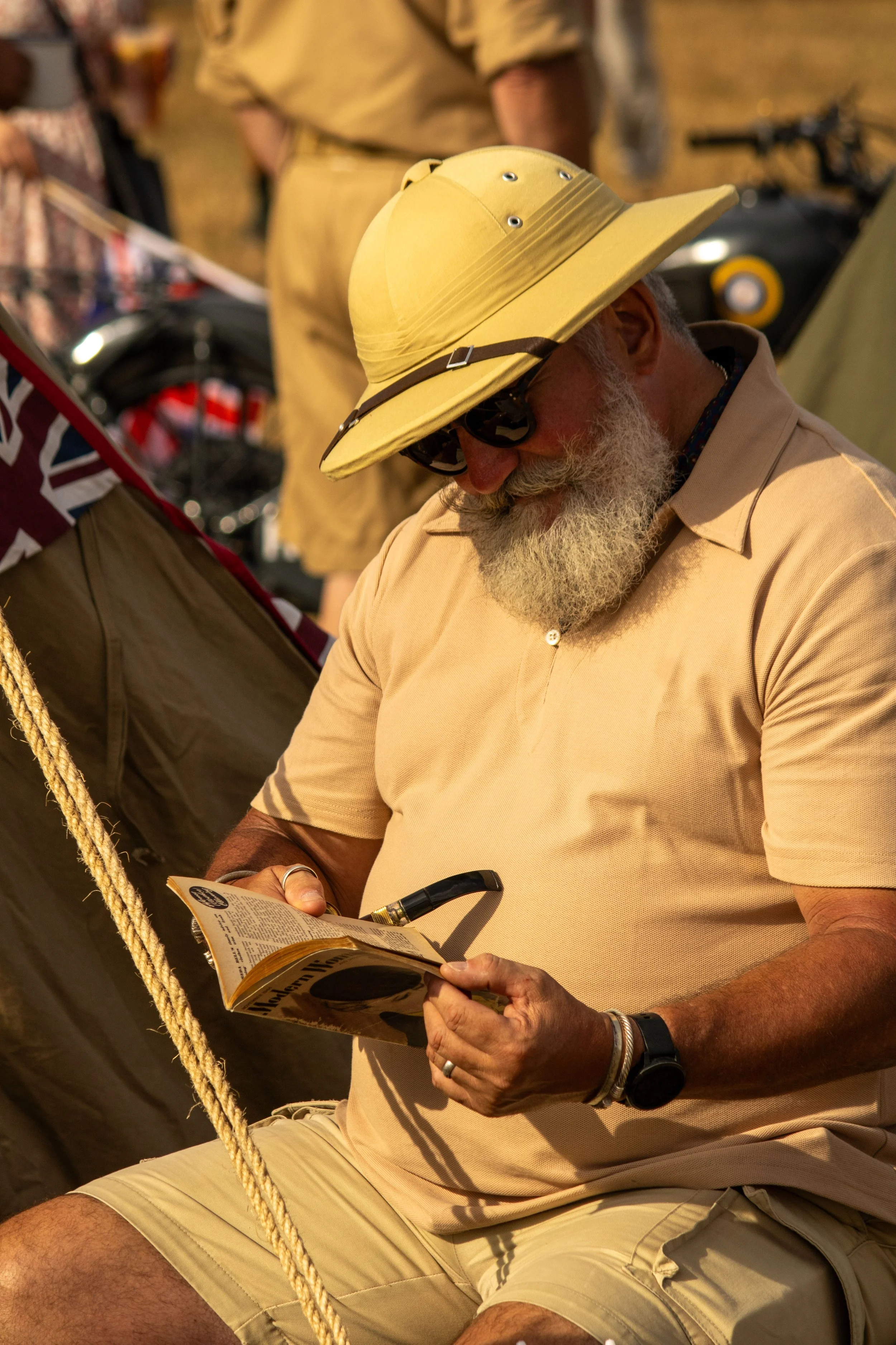 Older man with gray beard, wearing a large yellow wide-brimmed hat and sunglasses, sitting outdoors, reading a booklet, wearing beige polo shirt and shorts, with a black watch and bracelet, holding a pen.